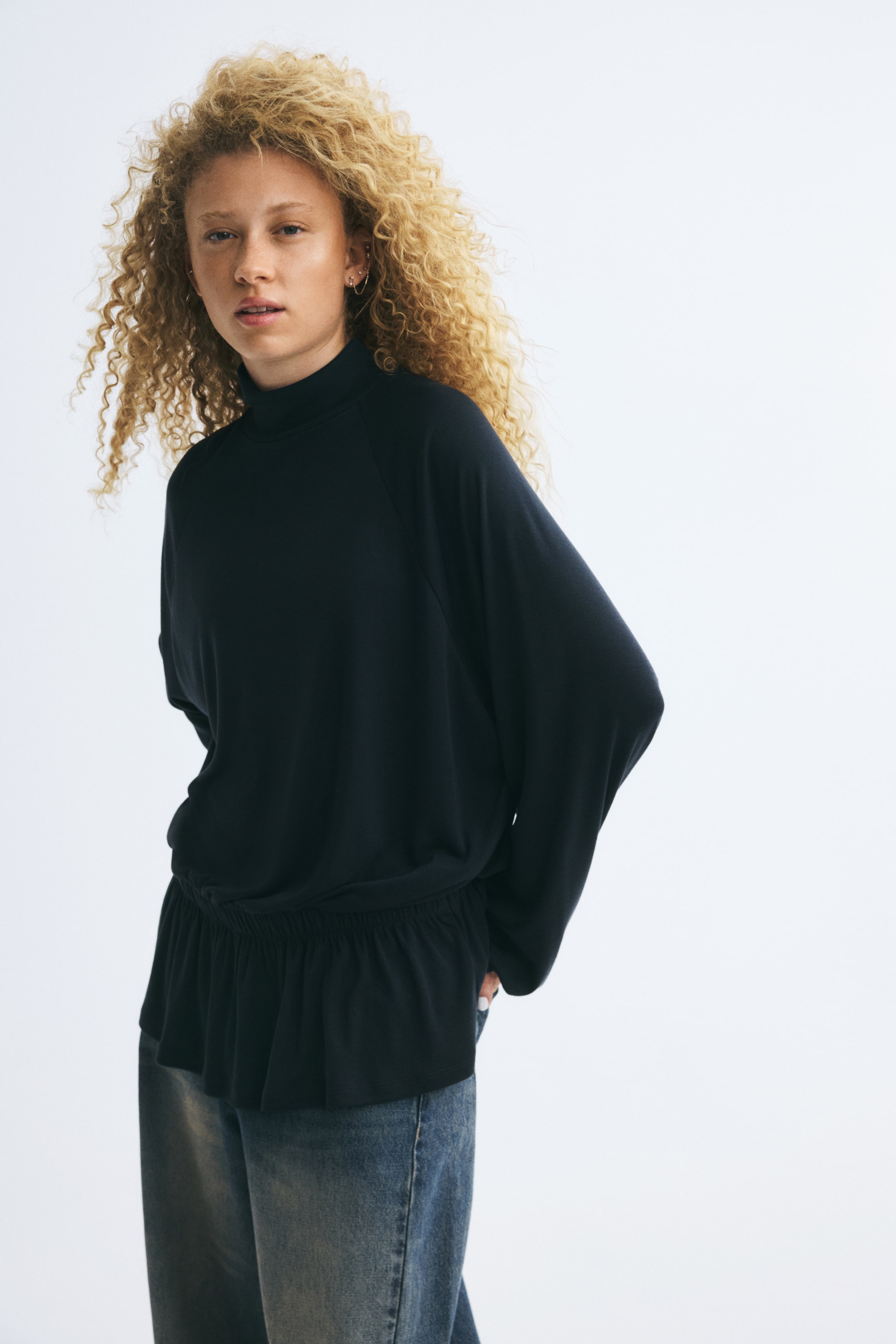 Ampliar la imagen: La modelo lleva puesta una blusa negra tipo jersey de corte oversized, con cuello alto tipo mock neck, mangas raglán largas y cintura elástica con fruncido tipo smock. La blusa se combina con jeans azules.