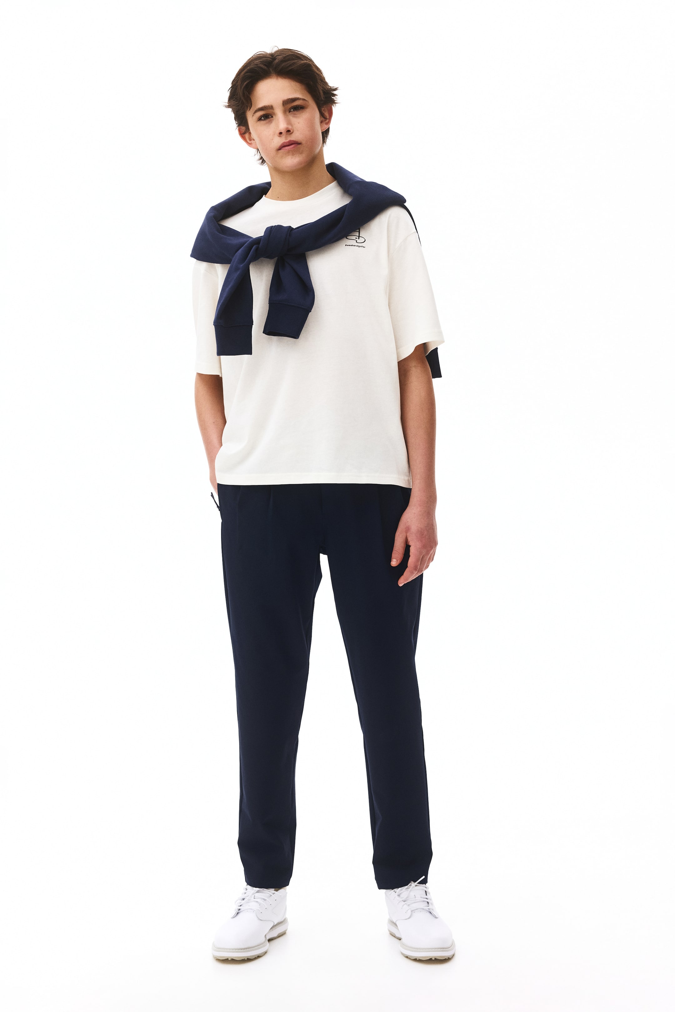 Ampliar la imagen: Una persona está modelando una camiseta de manga corta color crema, con un pequeño gráfico en el pecho izquierdo, y un suéter azul marino con puños acanalados atado alrededor de sus hombros. También lleva pantalones azul marino de corte relajado y tenis blancos con cordones, mirando hacia adelante.