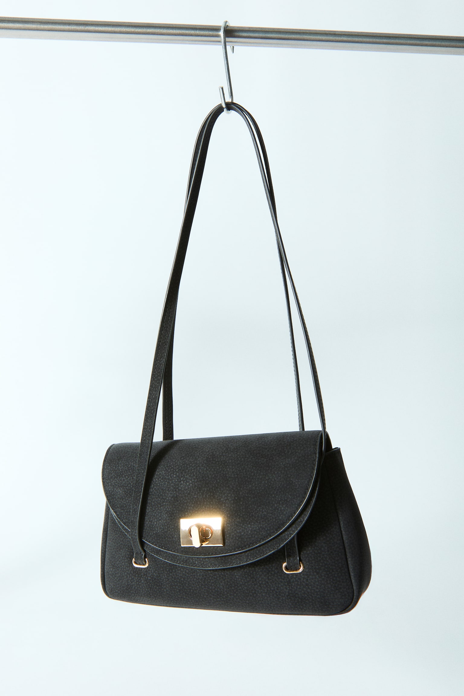 A black pebbled handbag is facing forward, featuring a curved flap closure with a gold-toned turn-lock. Two thin shoulder straps are extending upwards from the top edge. - 1