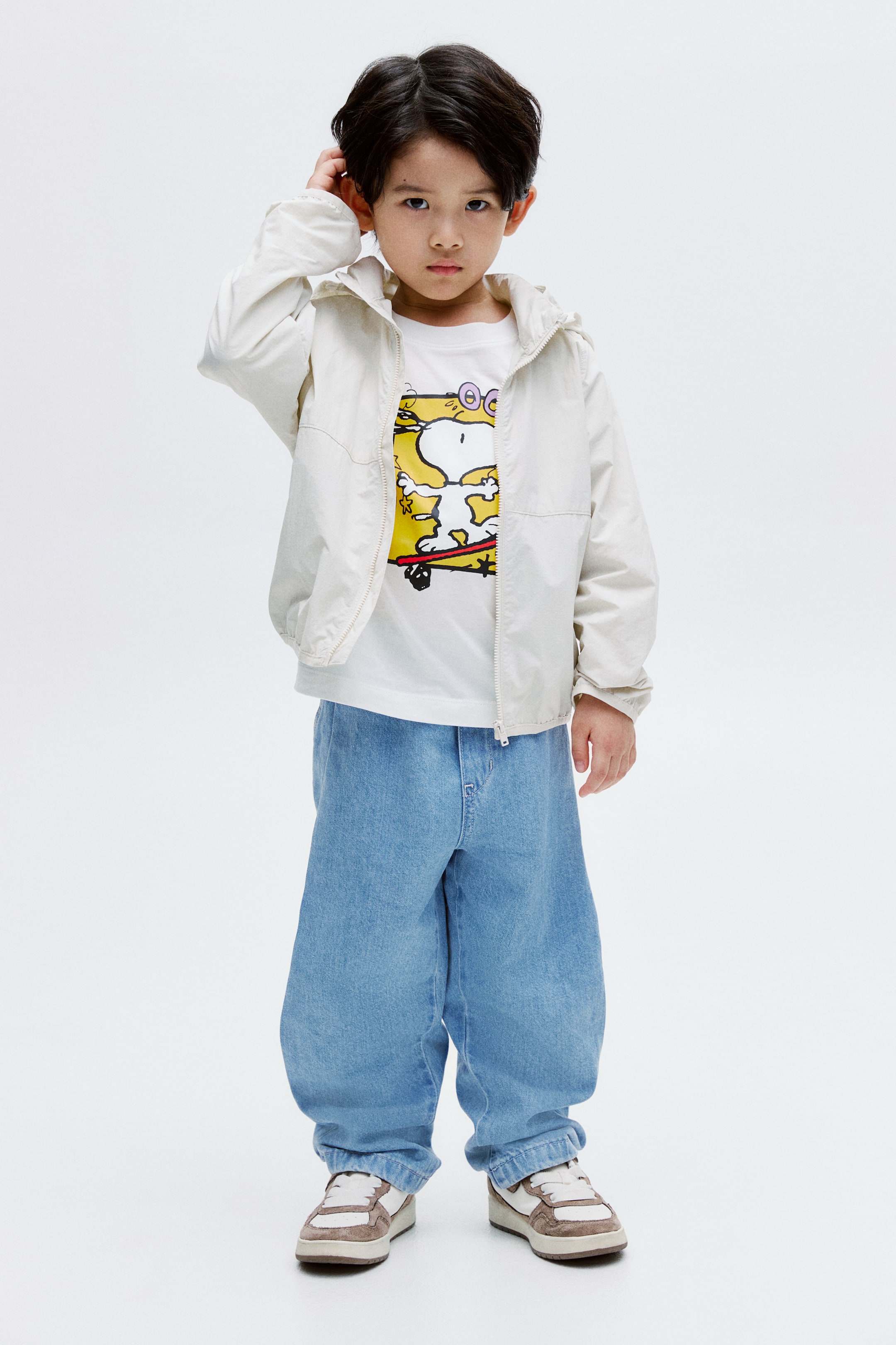 拡大画像を表示: 子供用のコーディネートは、ライトベージュのフード付きジャケットをオープンにして着用し、黄色のスヌーピーグラフィックが施された白いTシャツを見せています。ライトブルーのワイドレッグデニムジーンズは、足首を絞ったデザインで、白とブラウンのレースアップスニーカーと共にスタイルを完成させています。