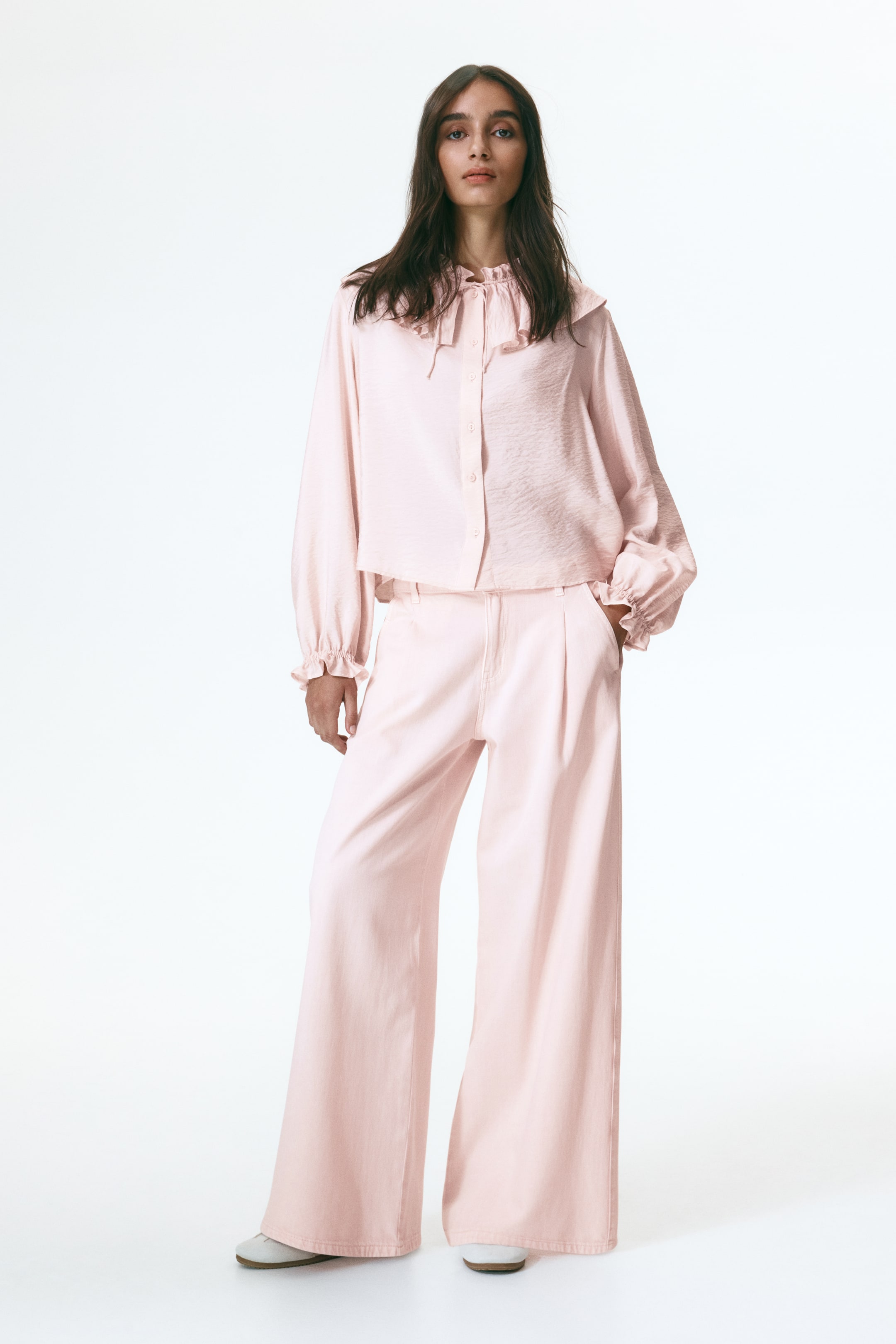 Ver imagem maior: Um conjunto rosa pálido apresenta uma blusa com folhos, gola alta, detalhes de laço, mangas compridas volumosas com punhos com folhos e uma frente com botões. É combinado com calças de perna larga a condizer, que exibem pregas frontais e bolsos laterais.