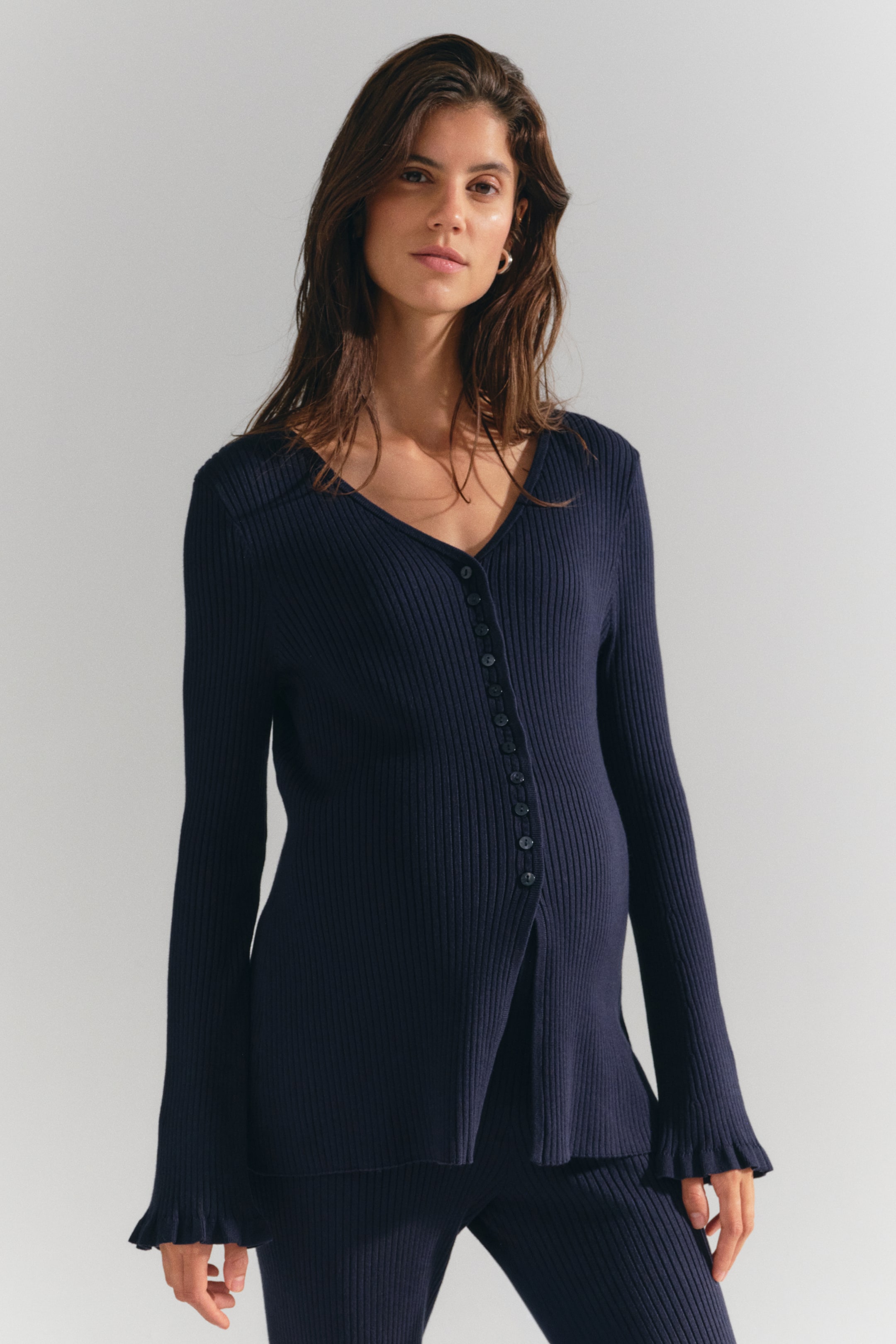 View larger image: A long, fitted, dark blue rib-knit cardigan is worn by the model, featuring a V-neckline, front button fastening, and gently flared sleeves with frill-trimmed cuffs. The cardigan is styled with matching dark blue rib-knit trousers.