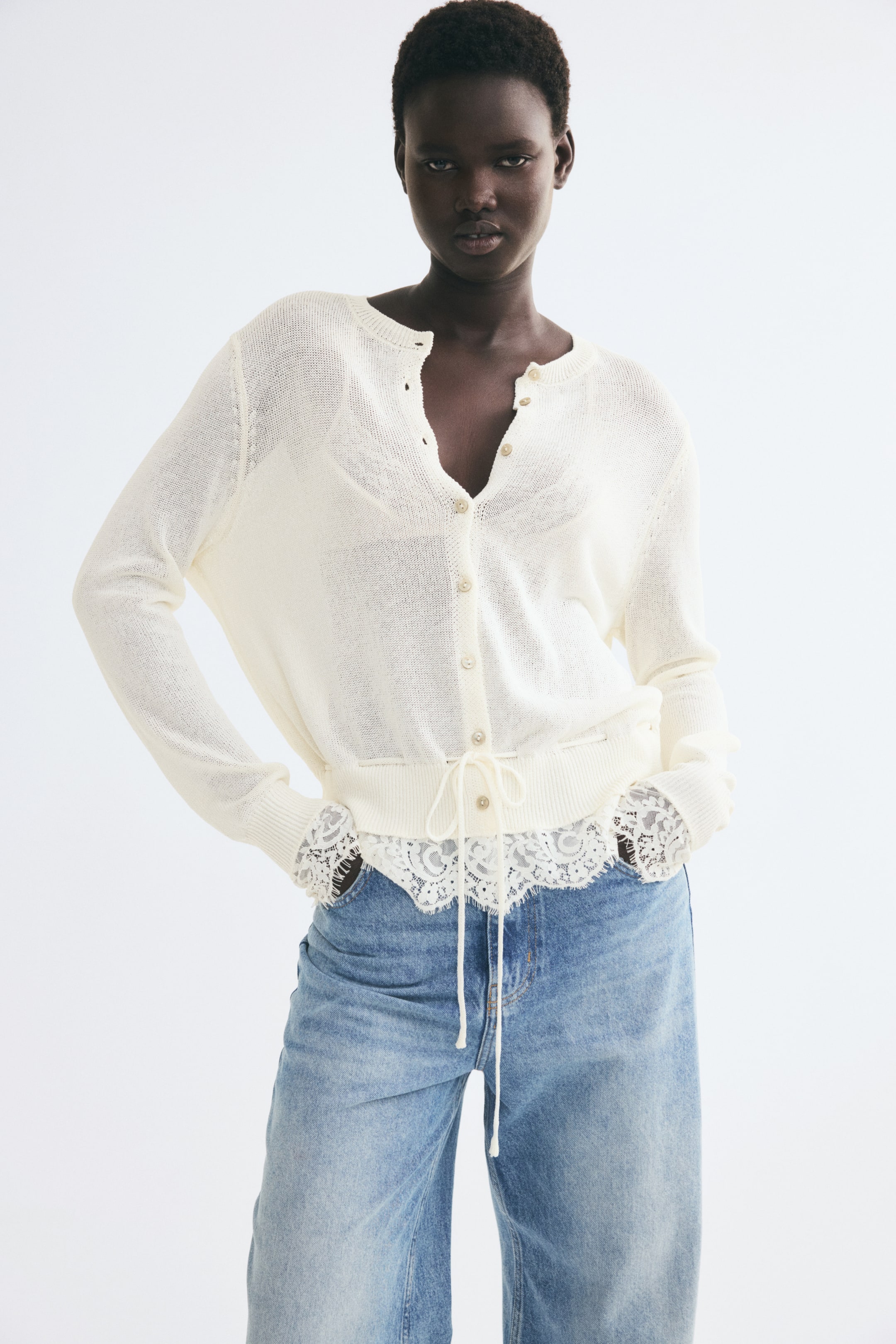 Ver imagem maior: A modelo está a usar um cardigan de malha fina em tom creme, combinado com jeans azul de lavagem clara. O cardigan apresenta fecho frontal com botões, mangas compridas com punhos canelados e acabamento em renda, e uma cintura canelada com cordão ajustável e acabamento em renda a estender-se abaixo da bainha.