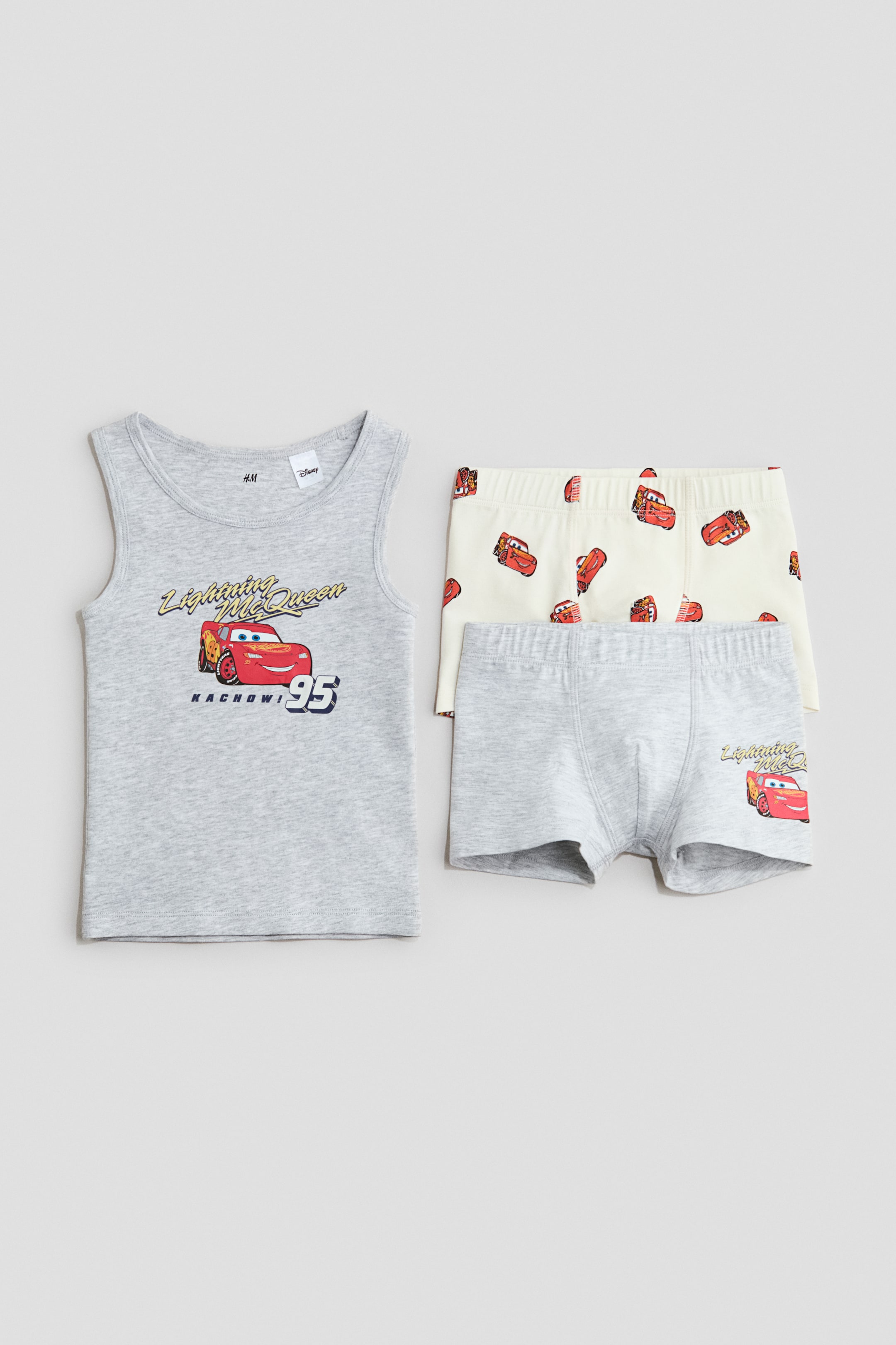Näytä suurempi kuva: Edestäpäin esillä on liivipaita ja kaksi paria shortseja. Harmaameleerattu liivipaita on varustettu ribbireunuksilla pääntiellä ja kädenteissä, sekä isolla etupainatuksella, jossa on Salama McQueen ja tekstit "Lightning McQueen" ja "Kachow! 95". Kahdessa shortsiparissa on joustavat vyötäröt; yksi kermanvärinen pari esittelee Salama McQueenin kokopainatusta, ja toinen harmaameleerattu pari sisältää pienemmän Salama McQueen -painatuksen edessä.