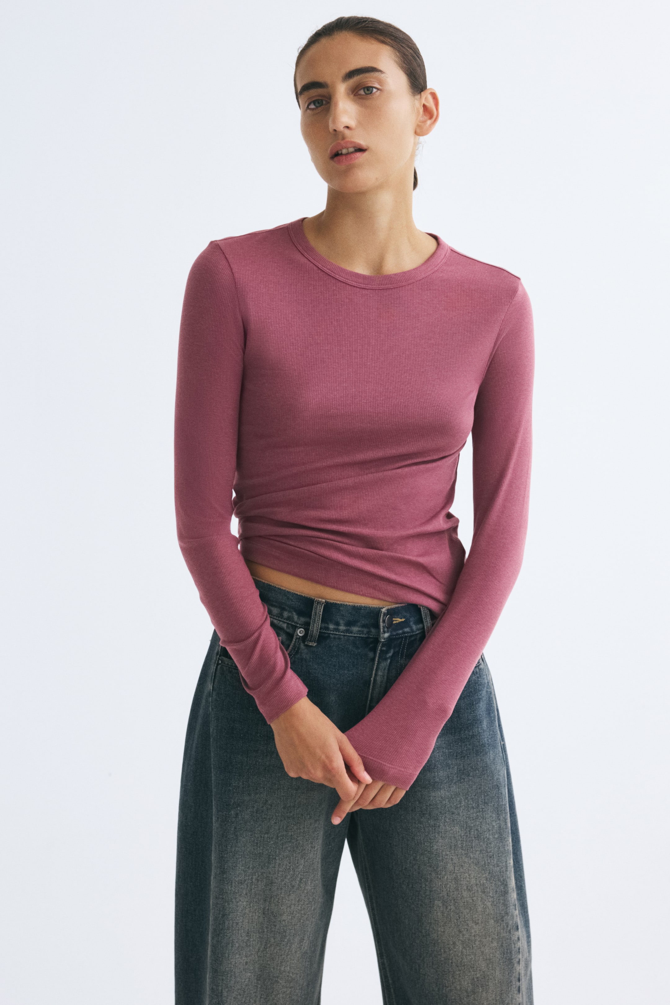 Voir une image plus grande: Le mannequin porte un haut ajusté en jersey côtelé rose poussiéreux, doté d’un col rond et de manches longues. Le haut est agencé avec un jean en denim à jambe large délavé foncé, tandis que le mannequin se tient debout, les mains croisées devant lui.