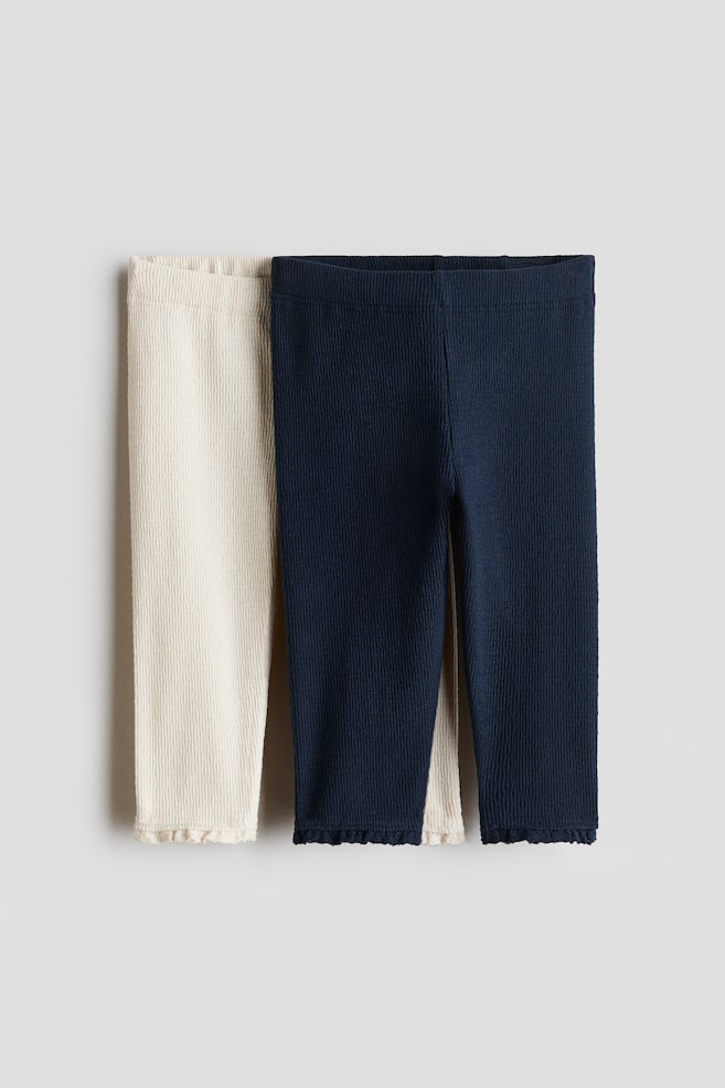 Ein Zweierpack gerippter Strickleggings ist von vorne zu sehen, wobei ein cremefarbenes Paar leicht hinter einem marineblauen Paar liegt. Beide Paare zeigen eine vertikale Rippstruktur, einen elastischen Bund und gewellte Lettuce-Edge-SÃ¤ume an den KnÃ¶chelbÃ¼ndchen. - 1