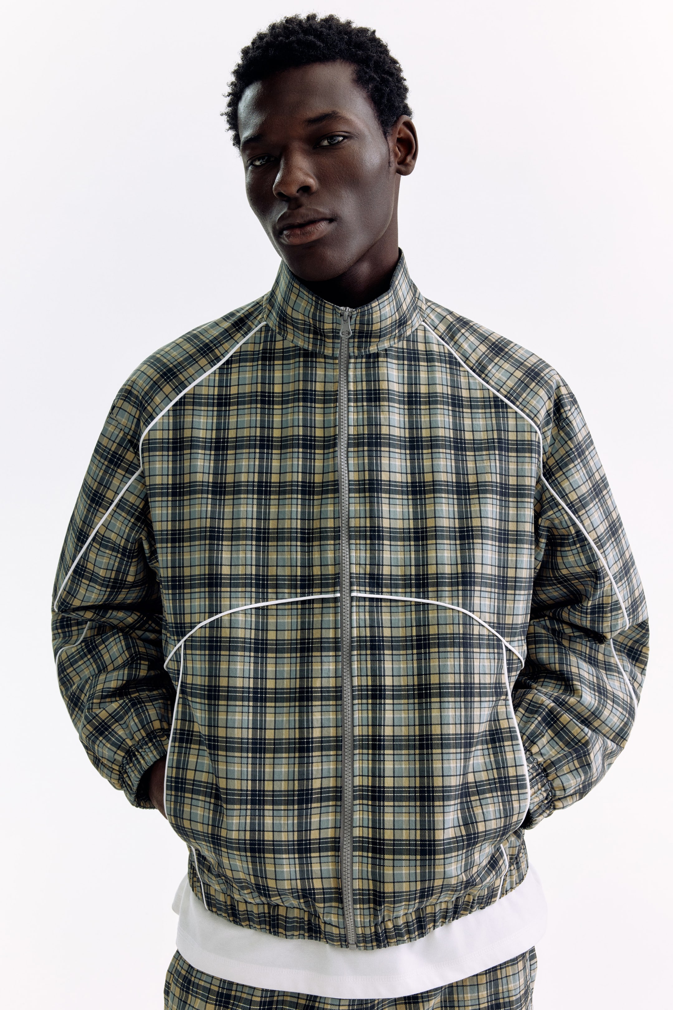 Ampliar la imagen: Una chaqueta de entrenamiento con cierre completo presenta un patrón de cuadros en tonos azul, verde y beige, con un cuello alto y un cierre frontal gris. Un ribete blanco delinea las mangas raglán y una línea horizontal a lo largo del pecho, con un dobladillo y puños elásticos.