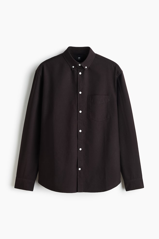 A dark brown Oxford cotton shirt is shown front on, showcasing a regular fit, a button-down collar, and a classic button-up front placket with contrasting white buttons. The shirt features a single open pocket on the left chest, long sleeves with buttoned cuffs, and a gently curved hem. - 1