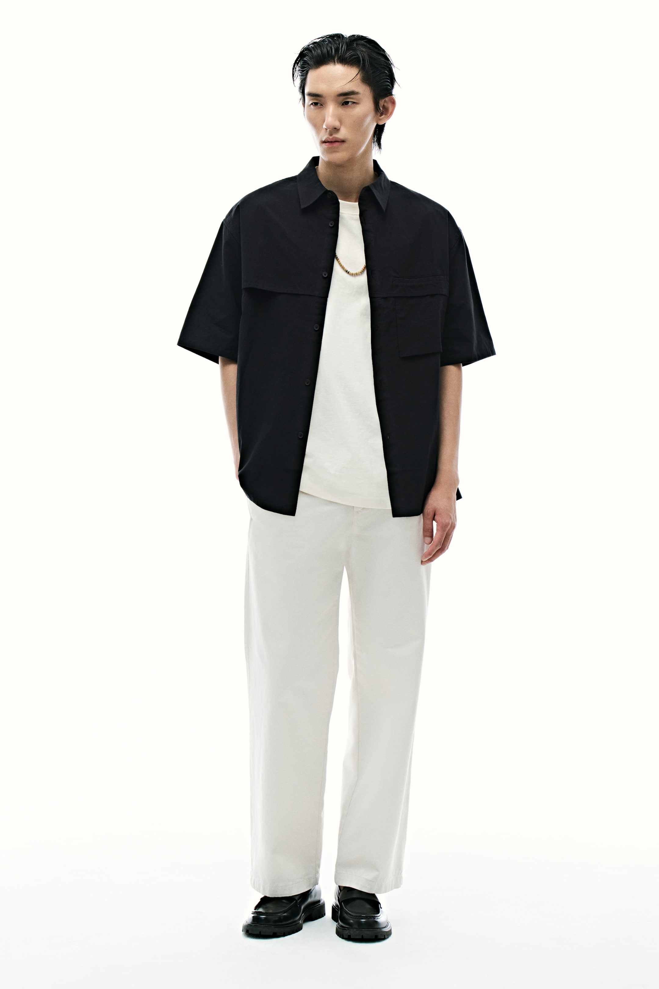 Xem ảnh lớn hơn: Người mẫu đang mặc một chiếc áo sơ mi đen bằng vải cotton twill không cài cúc bên ngoài áo thun trắng. Chiếc áo sơ mi này có cổ áo, tay ngắn, yếm trước rộng, chi tiết túi giả trên ngực trái, túi vá lộ ra trên ngực phải, và gấu áo cắt thẳng.