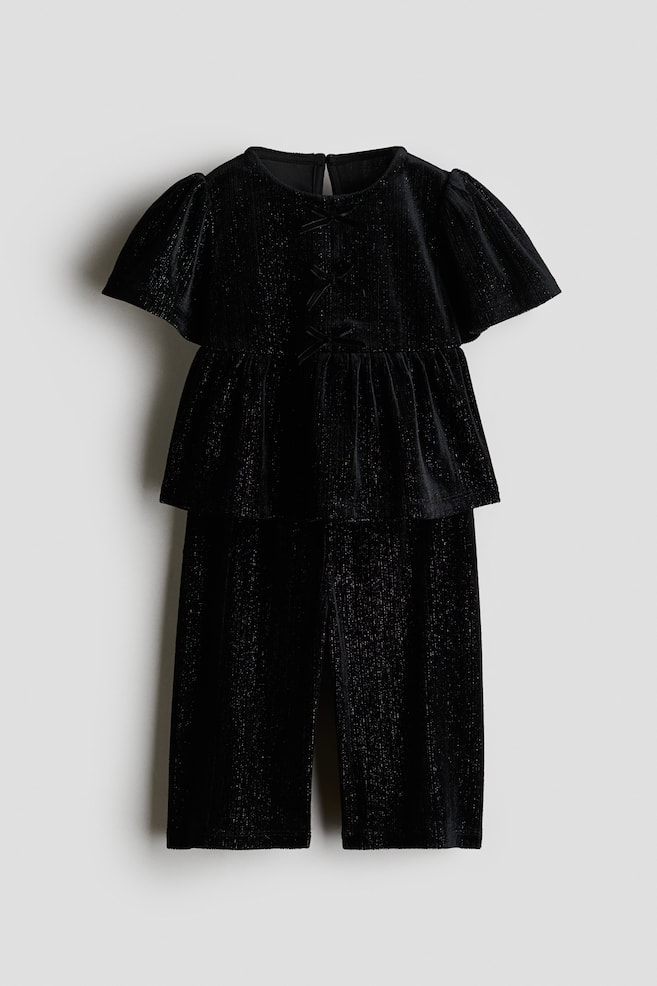 Een tweedelige zwarte kledingset, van voren weergegeven, bestaande uit een top met peplum en korte mouwen en een bijpassende broek met rechte pijpen, vervaardigd van een gestructureerde fluwelen stof met een subtiele glans. De top heeft een ronde halslijn, licht gepofte mouwen en drie kleine decoratieve strikjes langs het midden van het voorpand. - 1