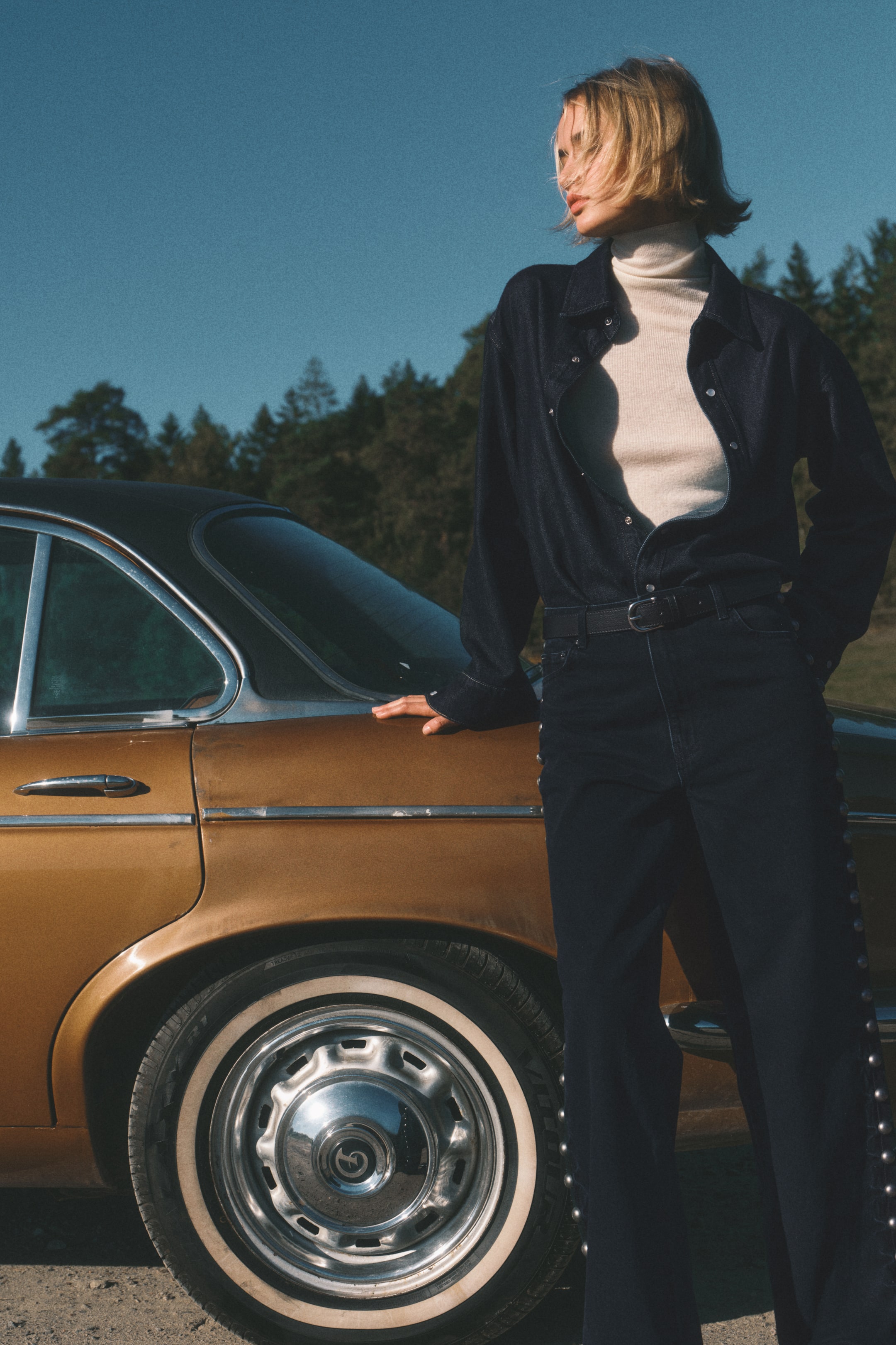 Voir une image plus grande: Une chemise en denim indigo foncé est portée ouverte, révélant un col roulé crème en dessous. Un jean indigo foncé assorti, avec une rangée de clous métalliques le long des coutures extérieures, est porté avec une ceinture noire, complétant la tenue.