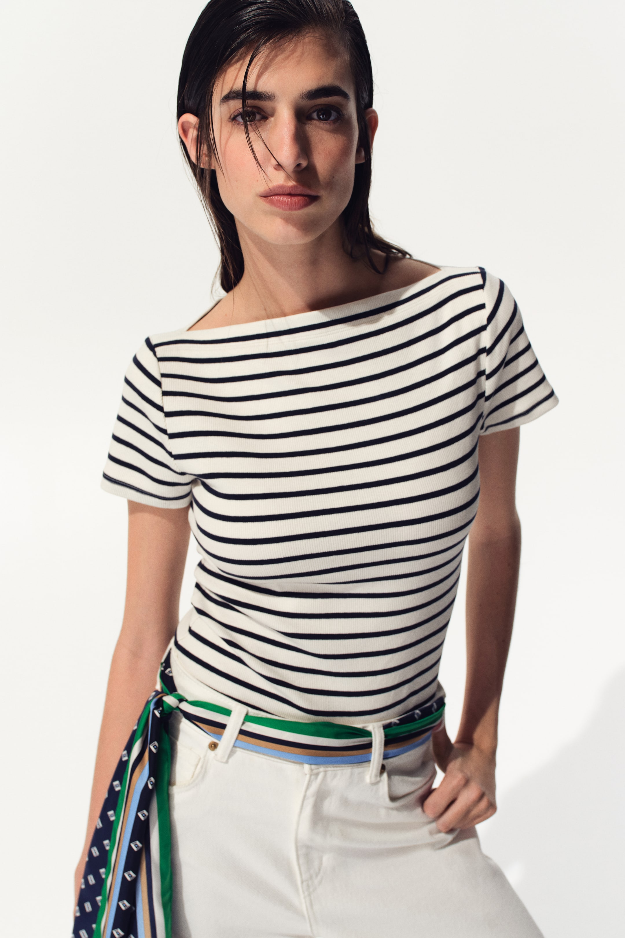View larger image: The model is wearing a fitted top made from ribbed cotton jersey, showcasing horizontal black and white stripes. It features a boat neckline and short sleeves. The top is paired with white pants and a patterned scarf tied around the waist.