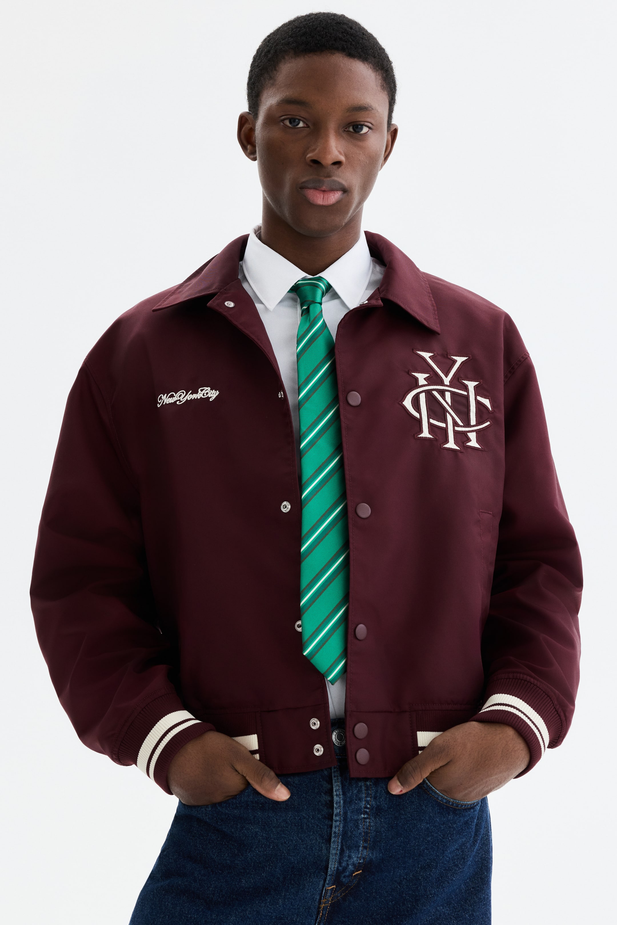 Ampliar la imagen: La chaqueta color marrón presenta un cuello en punta, un cierre frontal con botones a presión y puños y dobladillo acanalados con detalles de rayas blancas. Muestra "New York City" en bordado blanco en el lado izquierdo del pecho y un monograma "NY" entrelazado en el lado derecho del pecho, llevándose abierta sobre una camisa blanca y una corbata de rayas verdes.