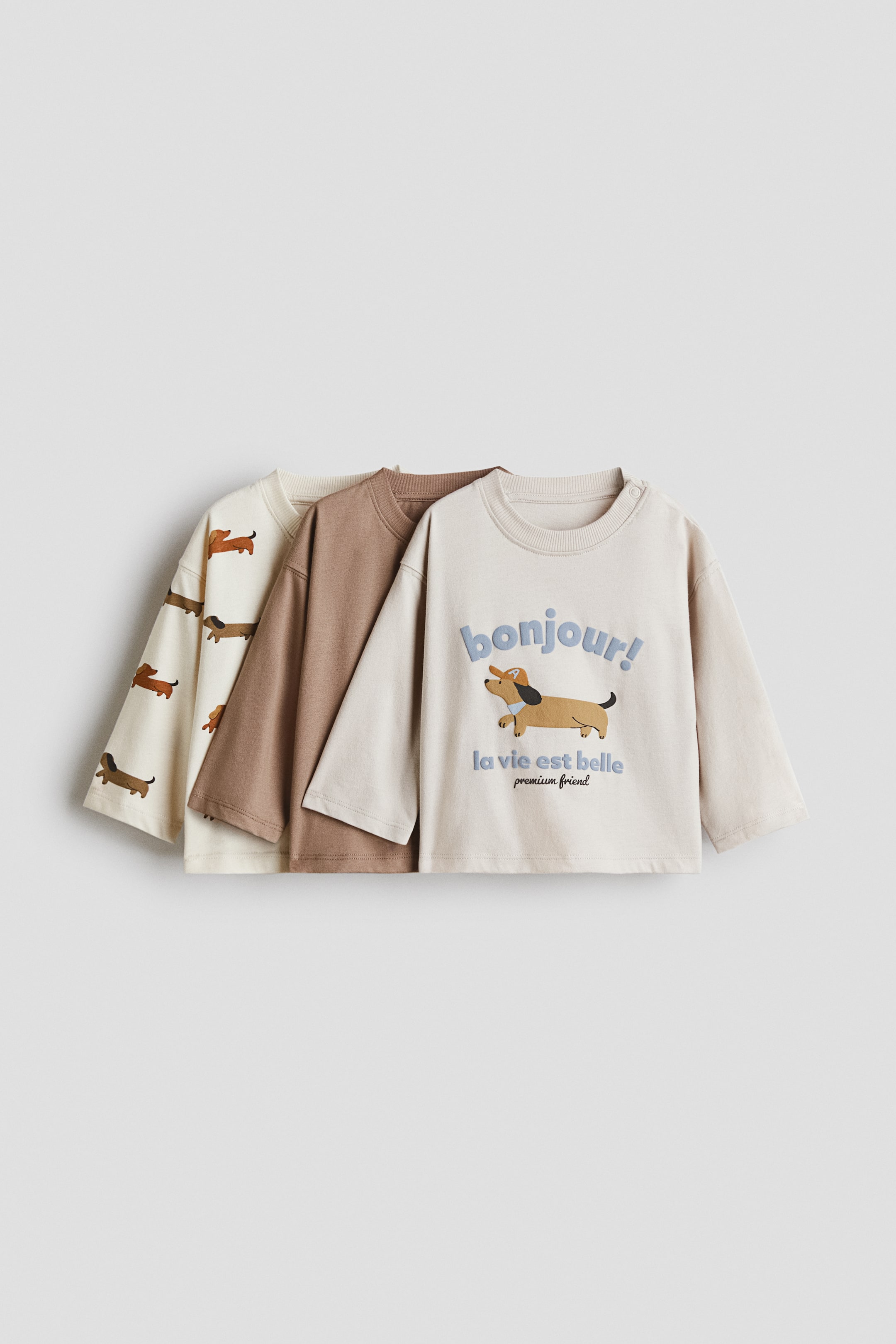 View larger image: A three-pack of long-sleeve cotton tees is displayed front-facing in a layered arrangement, showcasing three distinct styles. The set includes an off-white tee with an all-over dachshund pattern, a solid brown tee, and a light beige tee featuring a central dachshund graphic with blue text reading “bonjour!” and “la vie est belle”.