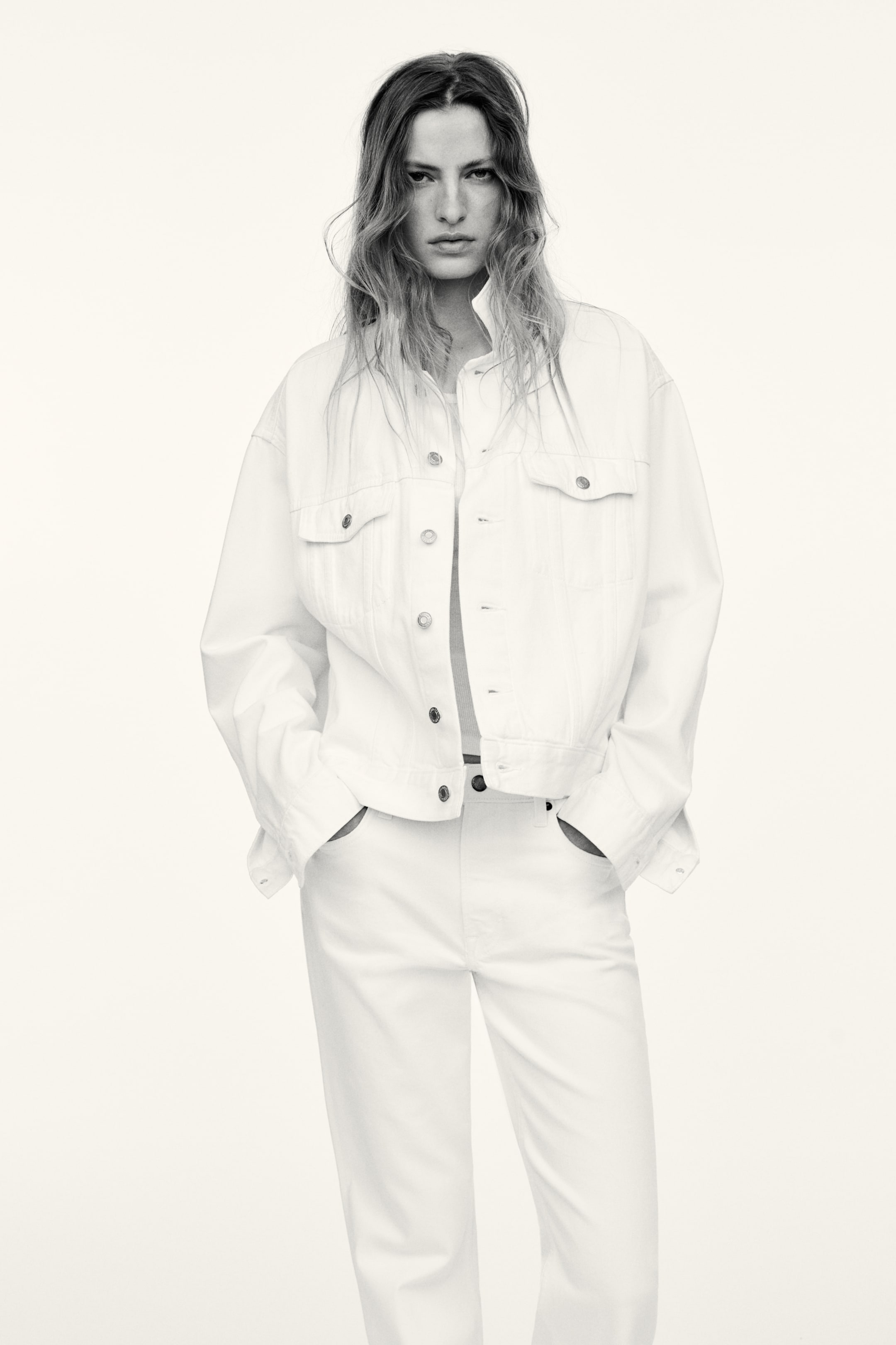 View larger image: "A white denim jacket is being worn unbuttoned, showcasing a pointed collar, two chest flap pockets with button closures, and silver-toned buttons down the front. It is layered over matching white pants, which display visible side pockets as hands are casually resting in them."