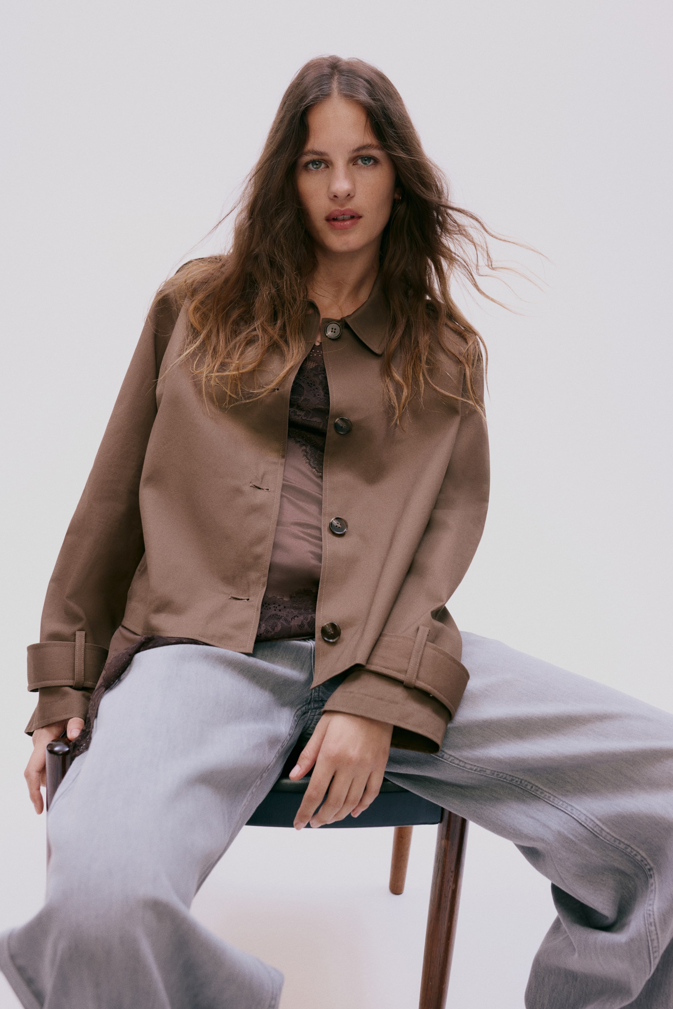 View larger image: A brown jacket features a pointed collar and a button-front closure with a few buttons undone. Its long sleeves display rolled-up cuffs secured by buttoned tabs, and it is worn open over a dark, sheer, lacy top.