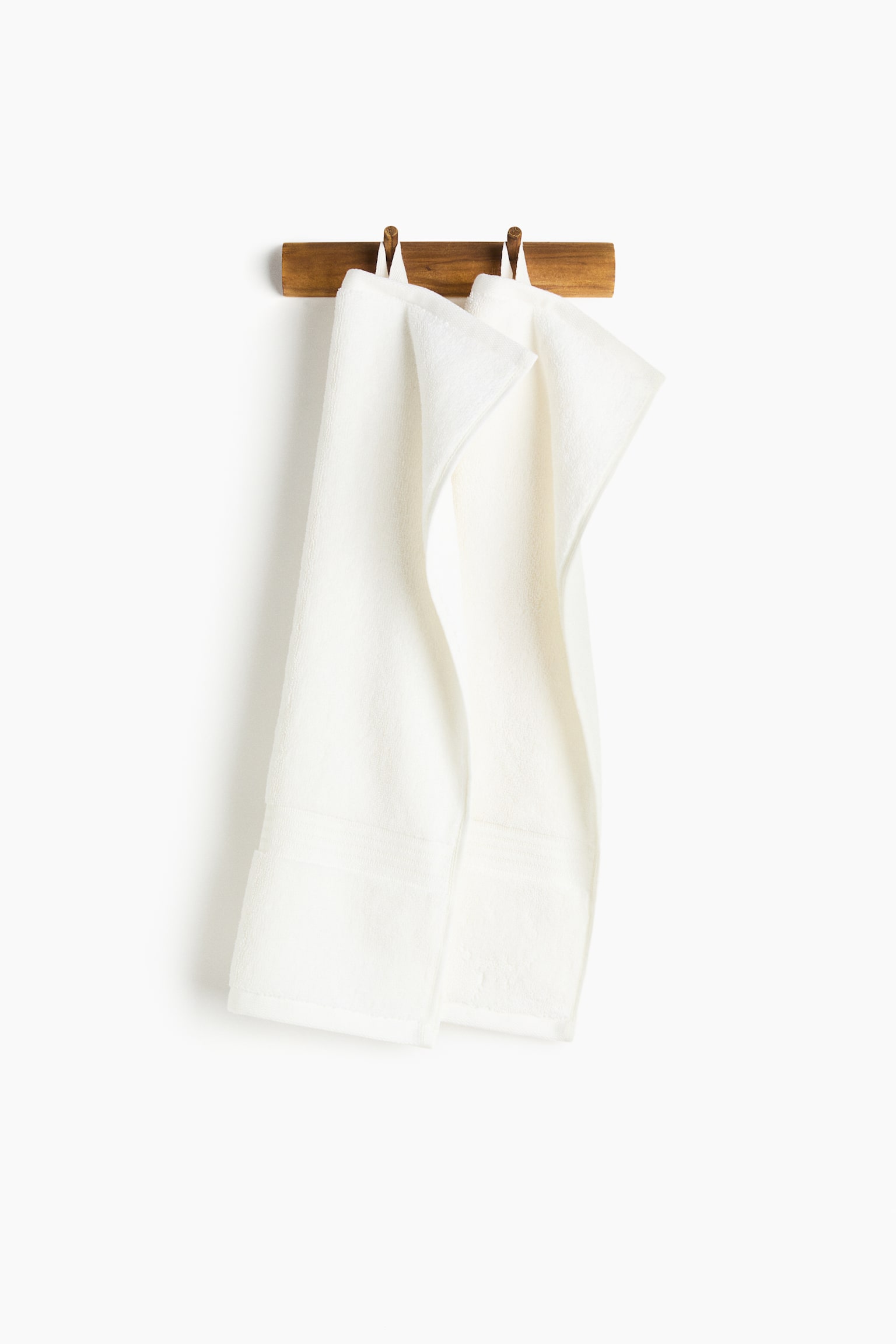 Two white terry cloth towels are hanging, folded over, from a wooden peg rack. Each towel is displaying a subtle horizontal band near its bottom edge. - 1