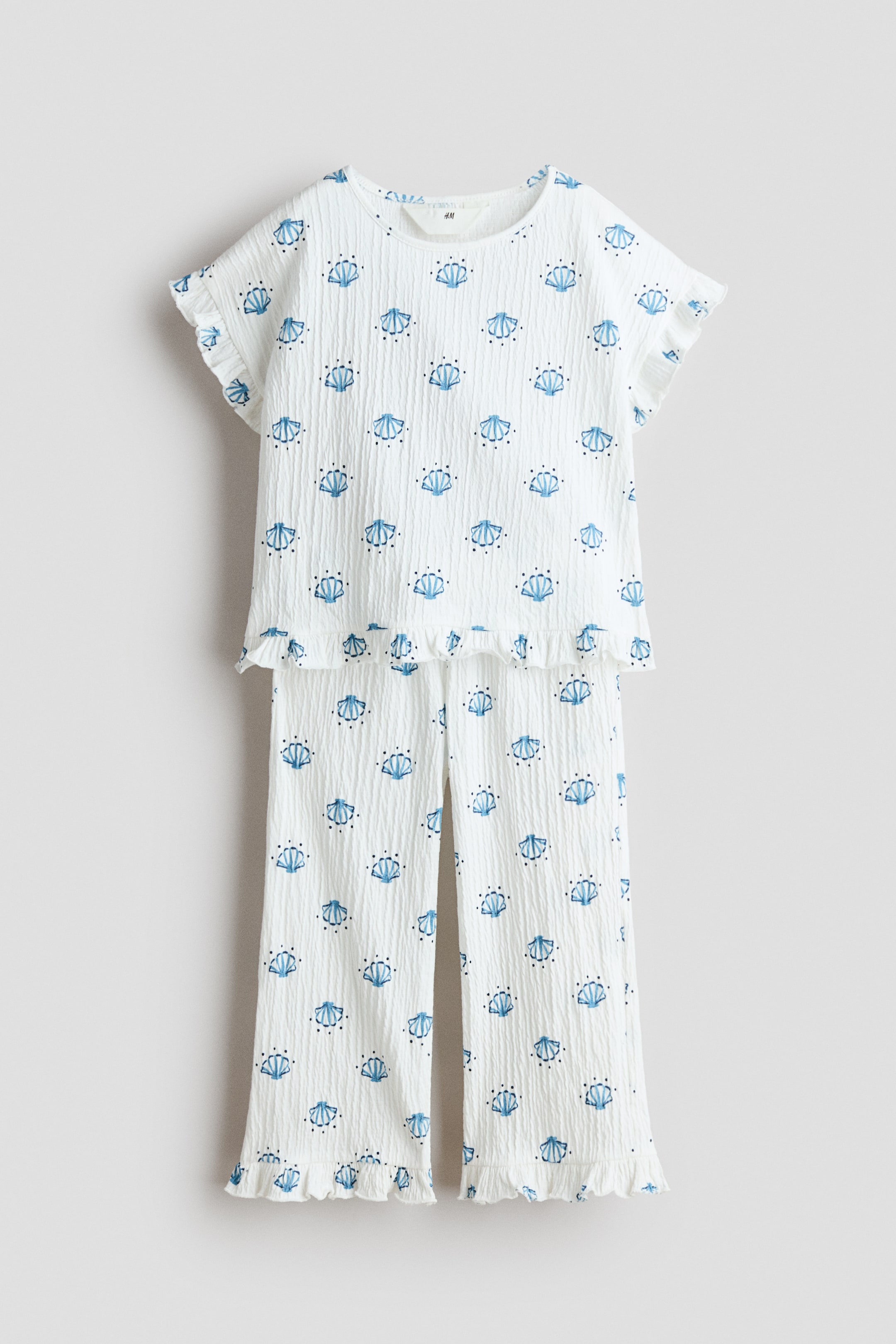 查看大圖: 這款兩件式睡衣套裝採用白色皺褶面料，搭配藍色海貝和點點圖案。前襟上衣設計為圓領、短荷葉袖及荷葉腰邊，而搭配的長褲則展示荷葉腳踝邊。