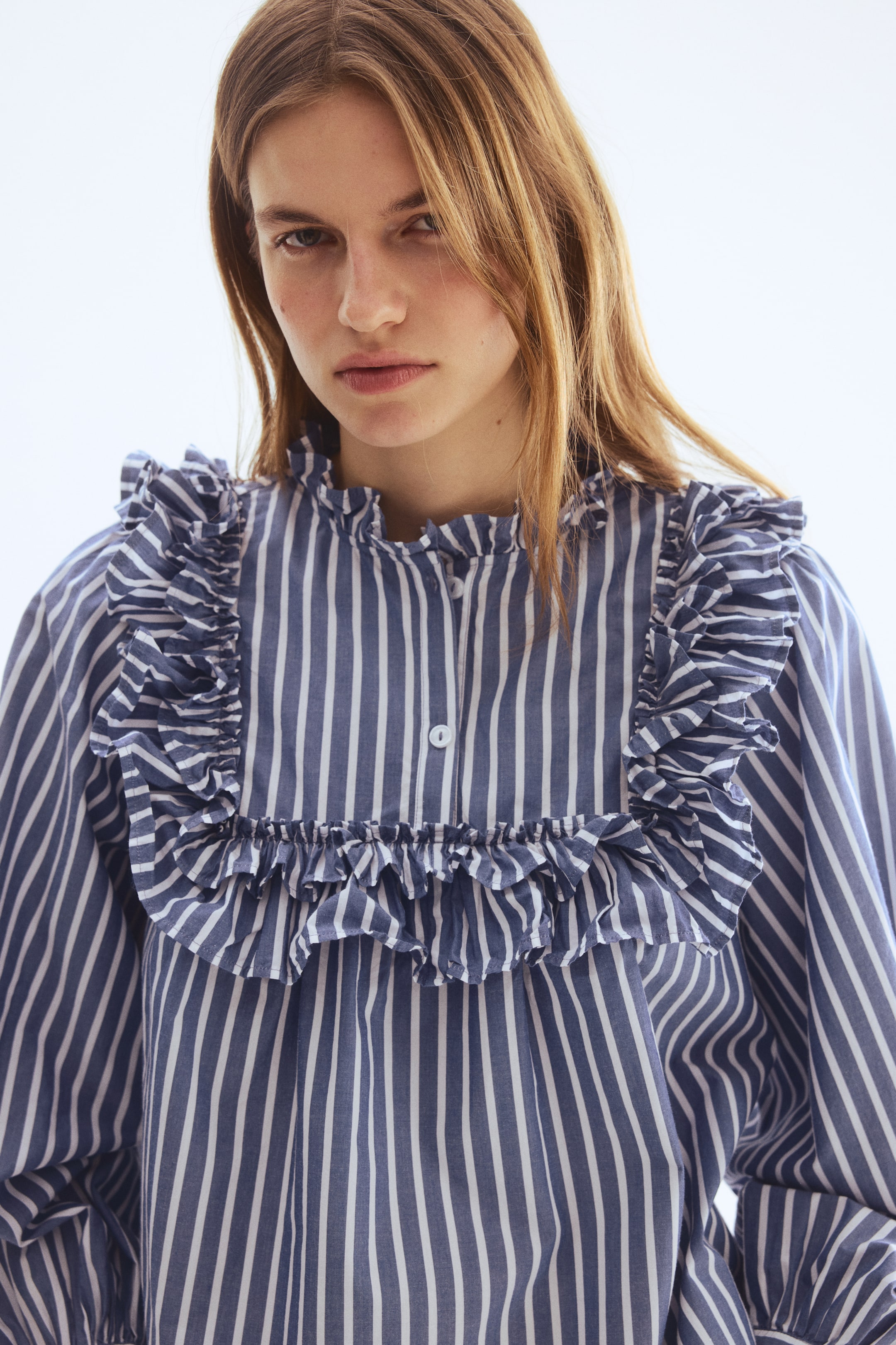 Ver imagen más grande: Una camisa de rayas azules y blancas presenta un cuello alto con volantes. Grandes volantes se extienden por el frente y sobre los hombros, y una tapeta parcial con botones blancos es visible en el centro del frente.