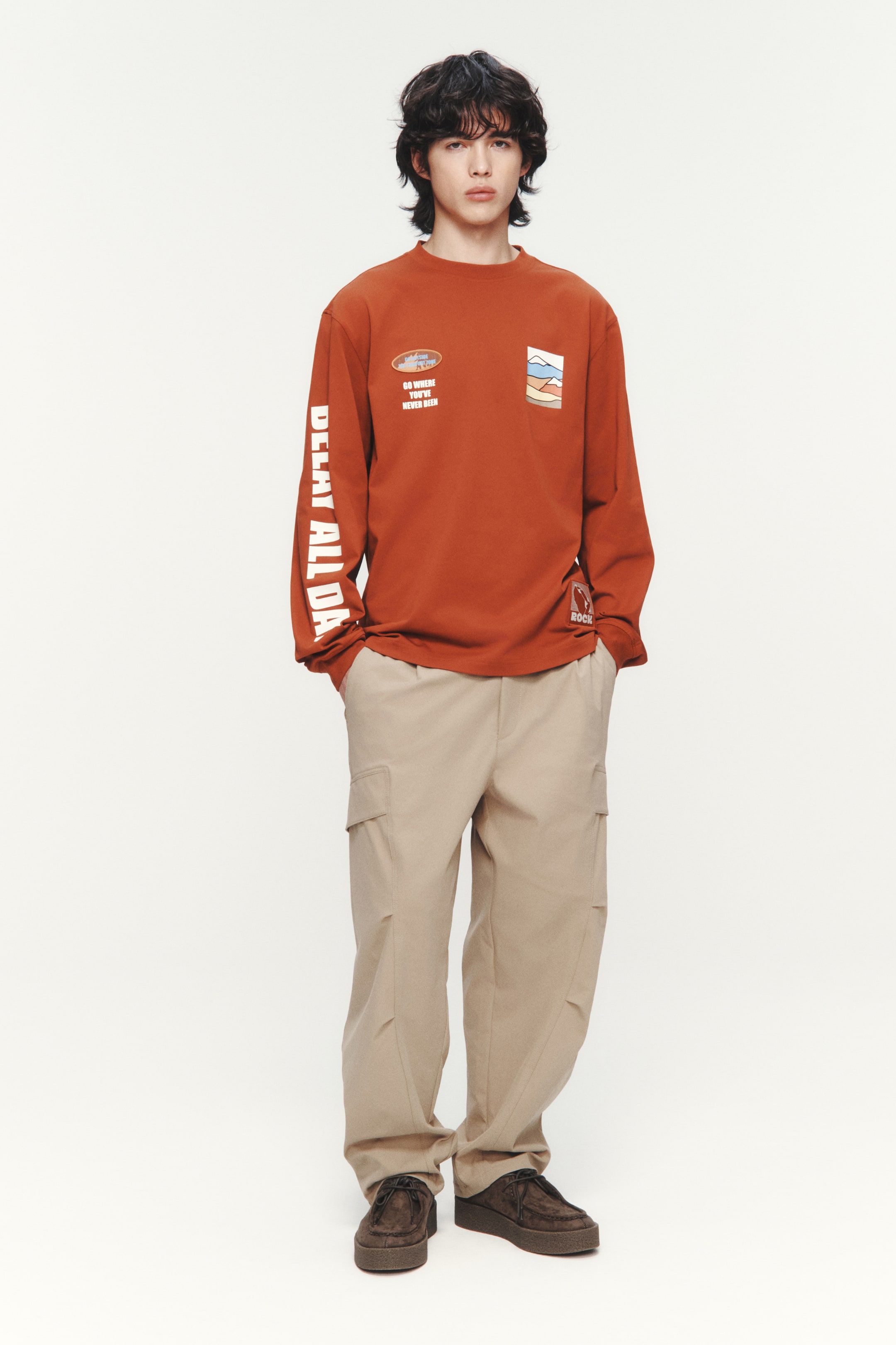 拡大画像を表示: モデルが着用しているのは、錆色のヘビーウェイトジャージートップで、ゆったりとしたフィット感を提供します。このトップは、ラウンドネックにリブトリム、ドロップショルダー、リブ付きカフス、ストレートカットの裾が特徴です。フロントと左袖にはテキストとグラフィックプリントが施されています。