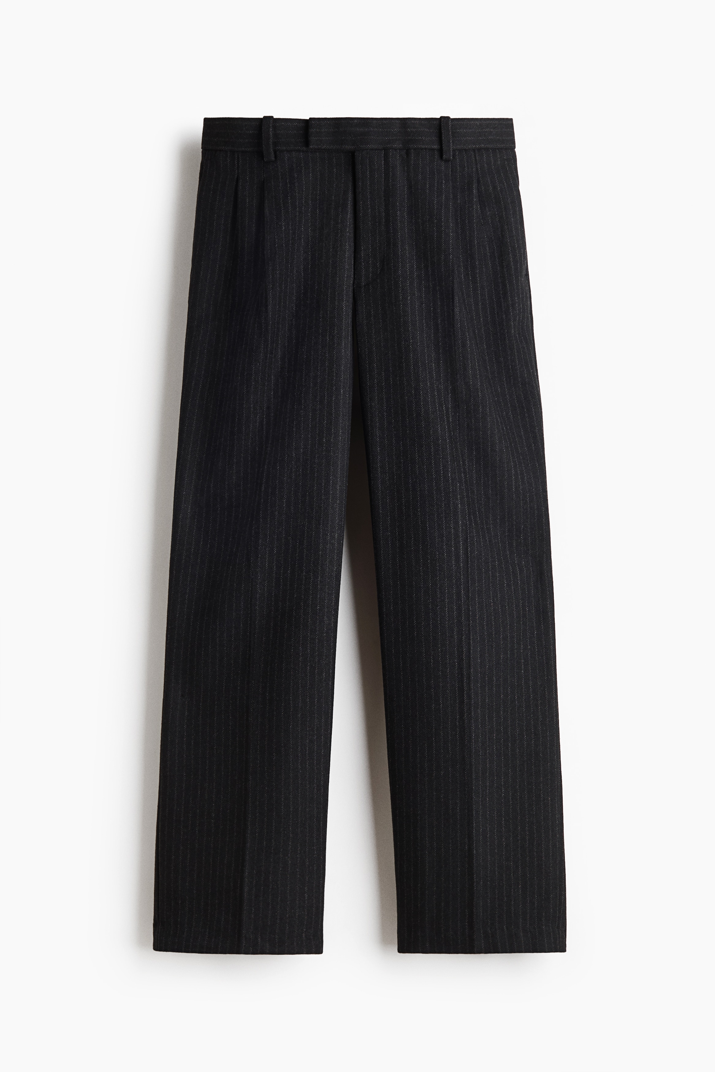 Le mannequin porte un pantalon sur mesure en flanelle à fines rayures foncées, avec une coupe décontractée, des jambes droites avec des plis marqués à l’avant et de subtils plis à la taille. Le pantalon est agencé avec un t-shirt blanc, un blouson zippé vert olive et des chaussures noires.