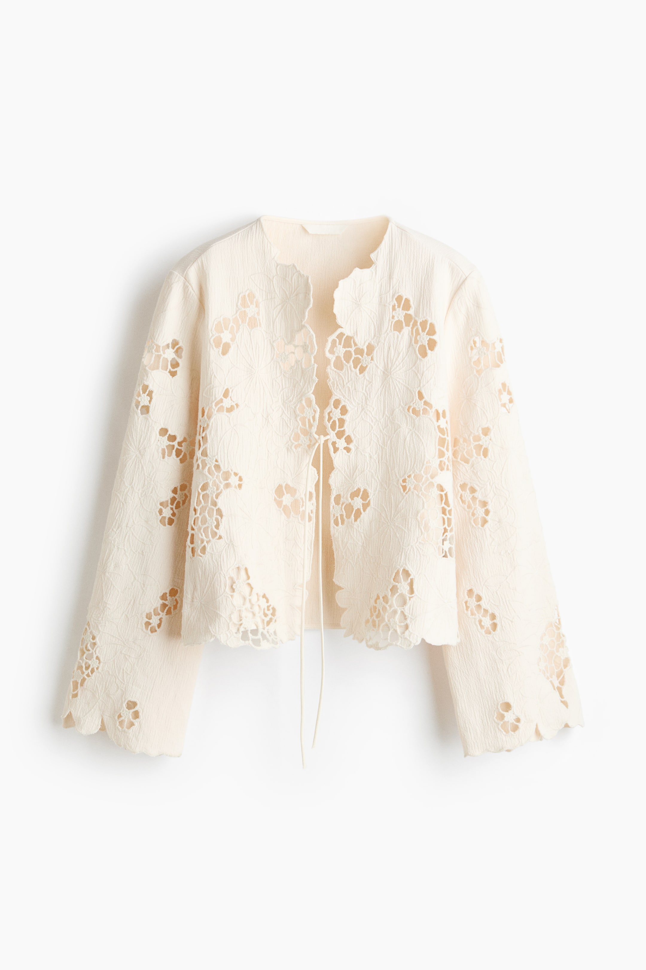 View larger image: A cream cropped jacket is showcased front-facing, highlighting an all-over cut-out floral design and scalloped edges along the neckline, front opening, hem, and wide cuffs. Two thin tie strings are visible at the front opening.