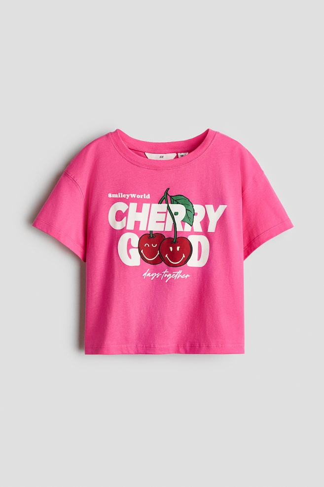Een roze T-shirt van katoenen jersey is van voren afgebeeld, met een ronde hals met ribboord en verlaagde schouders. Het shirt toont een grafische print op de voorkant met de tekst "CHERRY GOOD" in grote witte letters, waarbij twee rode kersen met glimlachende gezichtjes dienen als de letters "O". - 1