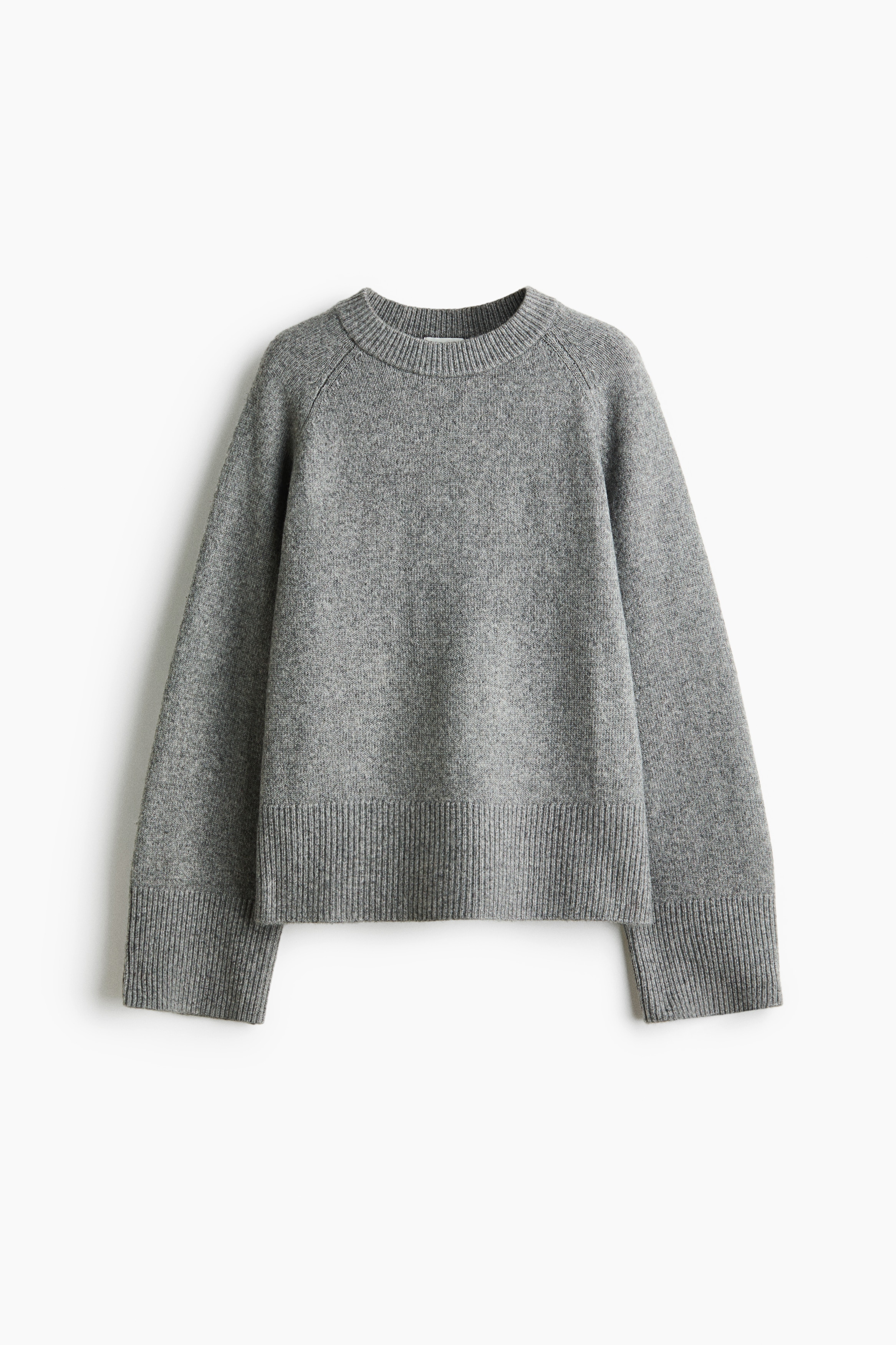 A grey, soft-knit jumper featuring a round, rib-trimmed neckline, long raglan sleeves, and wide ribbed cuffs is being worn by the model. The jumper is layered over a white long-sleeve shirt and paired with beige trousers.