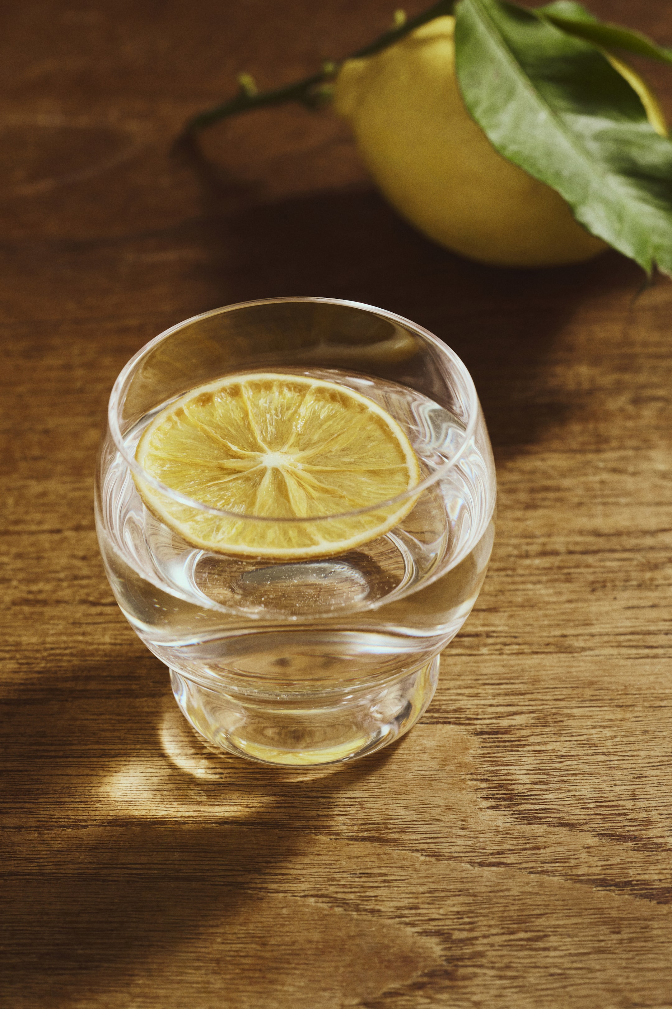 Voir une image plus grande: Un verre transparent contient un liquide clair, avec une tranche de citron jaune flottant à la surface. Le verre présente une forme arrondie et trapue, légèrement effilée au milieu, et est visible sous un angle légèrement surélevé.