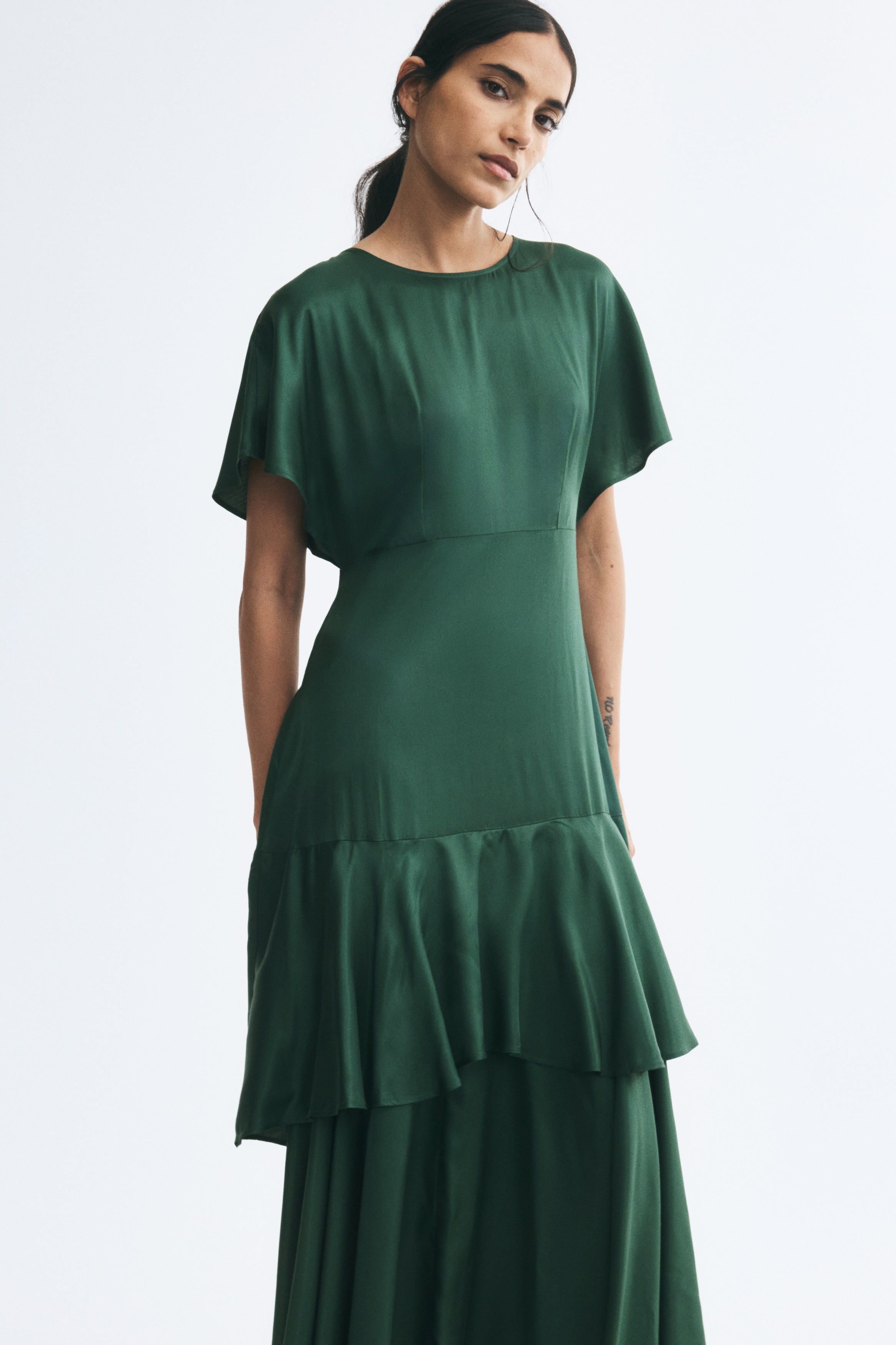 A dark green dress is shown from the front, featuring short flutter sleeves, a round neckline, and a defined waist. The dress cascades into a tiered, layered skirt with ruffled detailing and an asymmetrical hemline.