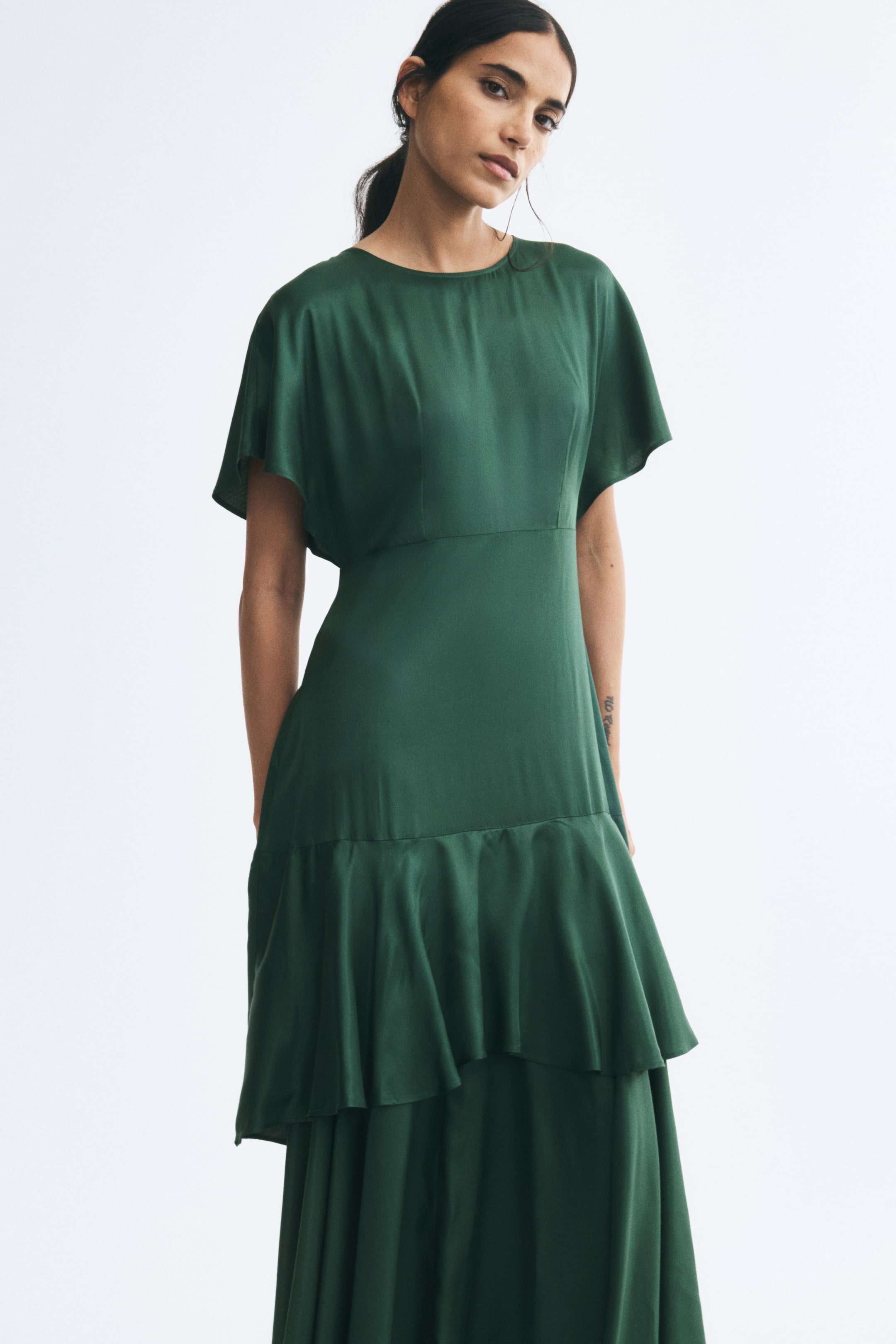 View larger image: The model is wearing a long green dress with a round neckline and short butterfly sleeves. The dress features a defined seam at the waist and a tiered skirt with a flounce below the hips.