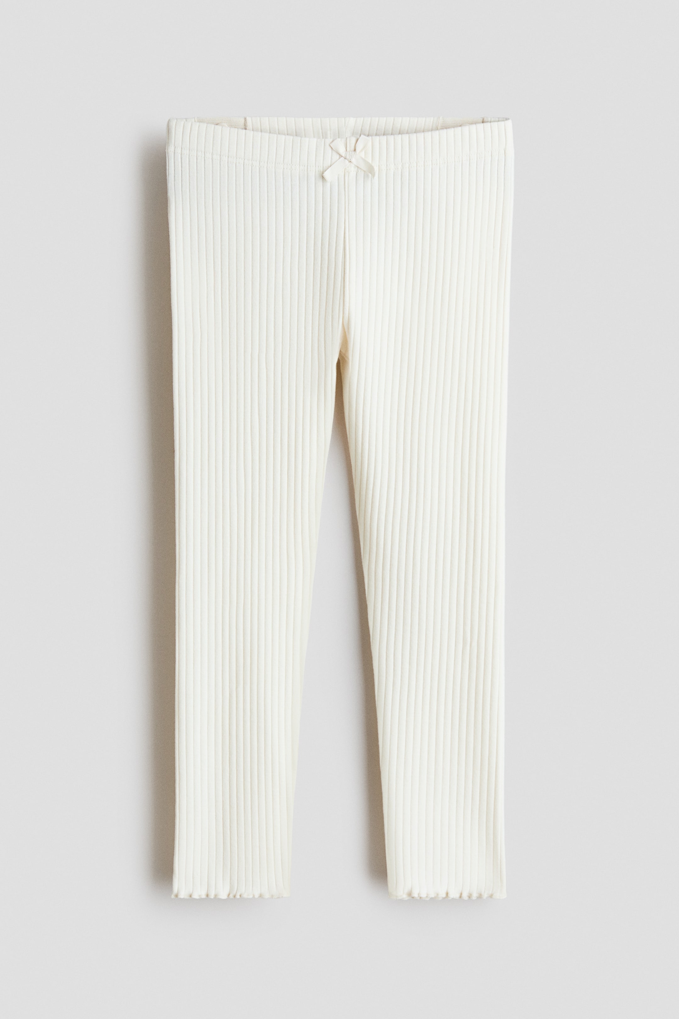 Ver imagem maior: Um par de leggings caneladas de cor creme é apresentado de frente. A cintura elástica apresenta um pequeno laço de cetim atado no centro frontal, e as aberturas das pernas têm acabamento com folhos.