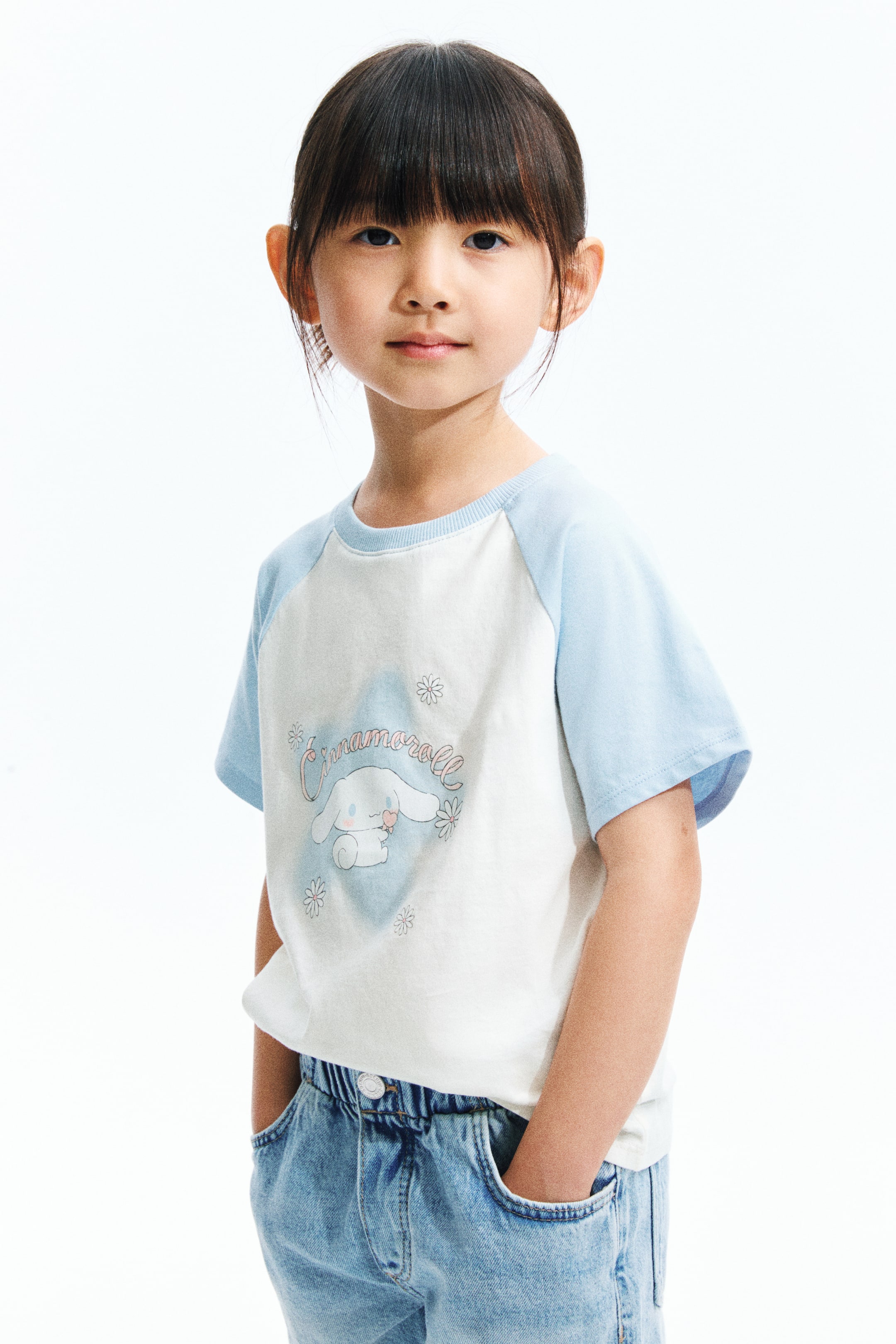 拡大画像を表示: 白いTシャツには、ライトブルーのラグランスリーブ、同色のリブ付きクルーネック、中央にはピンクのハートを持つ白いキャラクターのグラフィックが描かれています。その周りにはライトブルーの陰影、『シナモロール』の文字、小さな白い花があしらわれています。Tシャツはゆったりとしたフィット感で、ライトウォッシュのデニムジーンズにタックインされています。