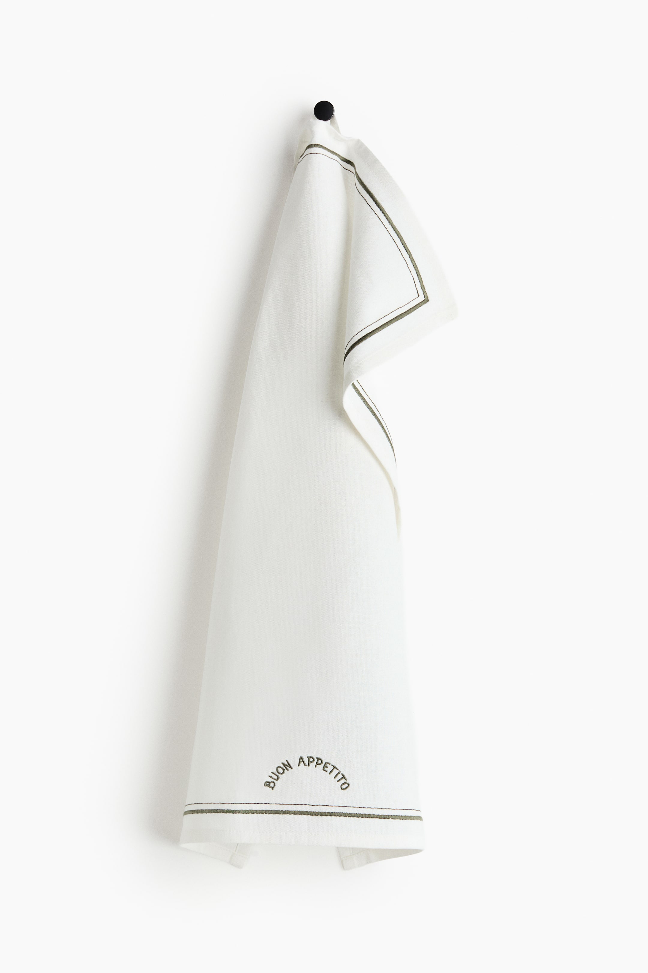 Преглед на по-голямо изображение: Бяла кухненска кърпа е закачена, прегъната през черна кука и представена отпред. По видимите ѝ ръбове се вижда тъмнозелена двойна линия, а в долната част е бродирано „BUON APPETITO“ в извита арка.