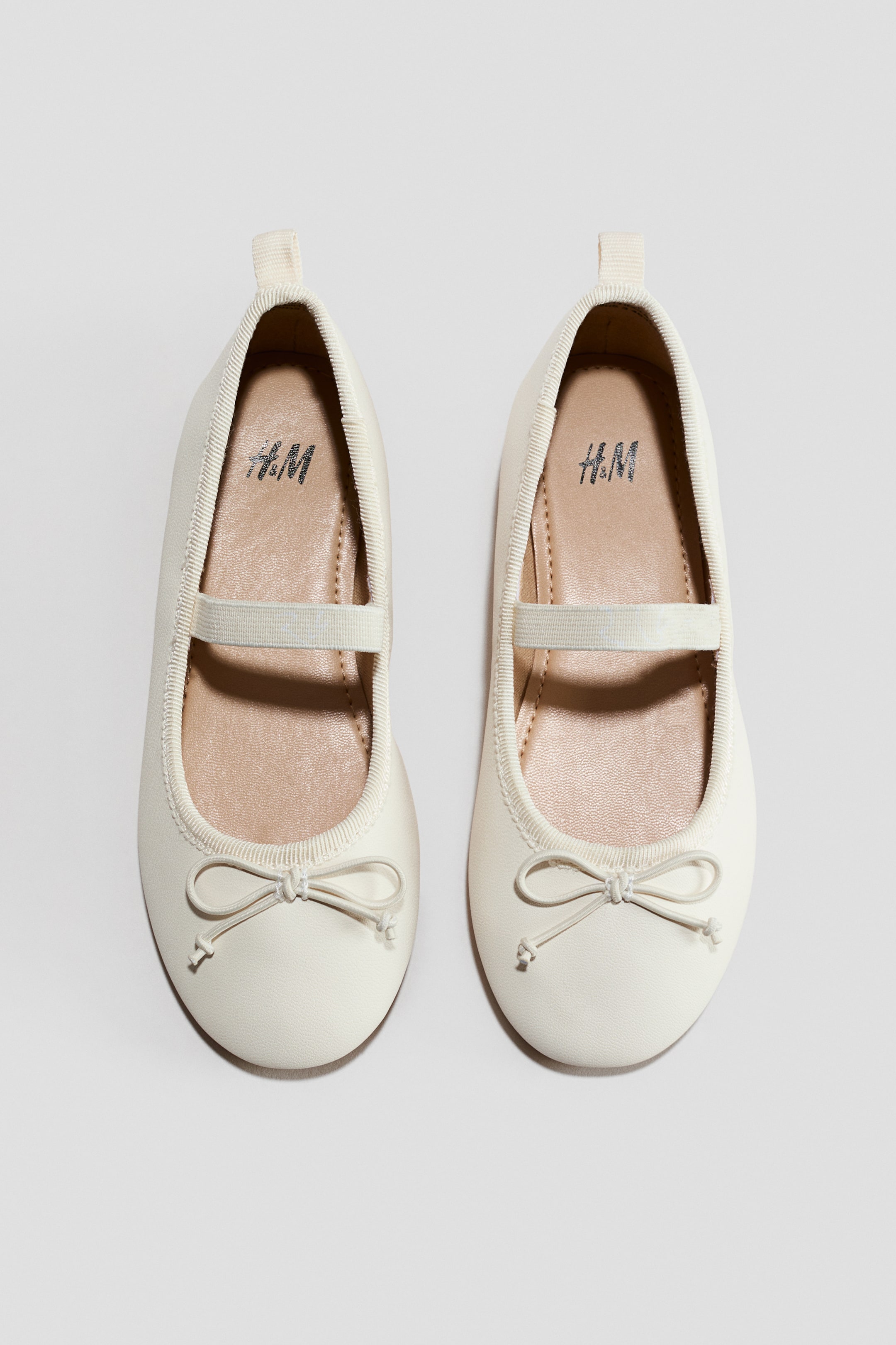 Voir une image plus grande: Une paire de ballerines crème présentée DE FACE, avec des bouts arrondis et un bord élastique nervuré autour de l'ouverture. Une lanière élastique traverse le dessus de chaque pied, et un nœud décoratif orne l'avant de chaque chaussure, tandis que les semelles intérieures beige clair affichent un logo argenté H&M visible à l'intérieur.