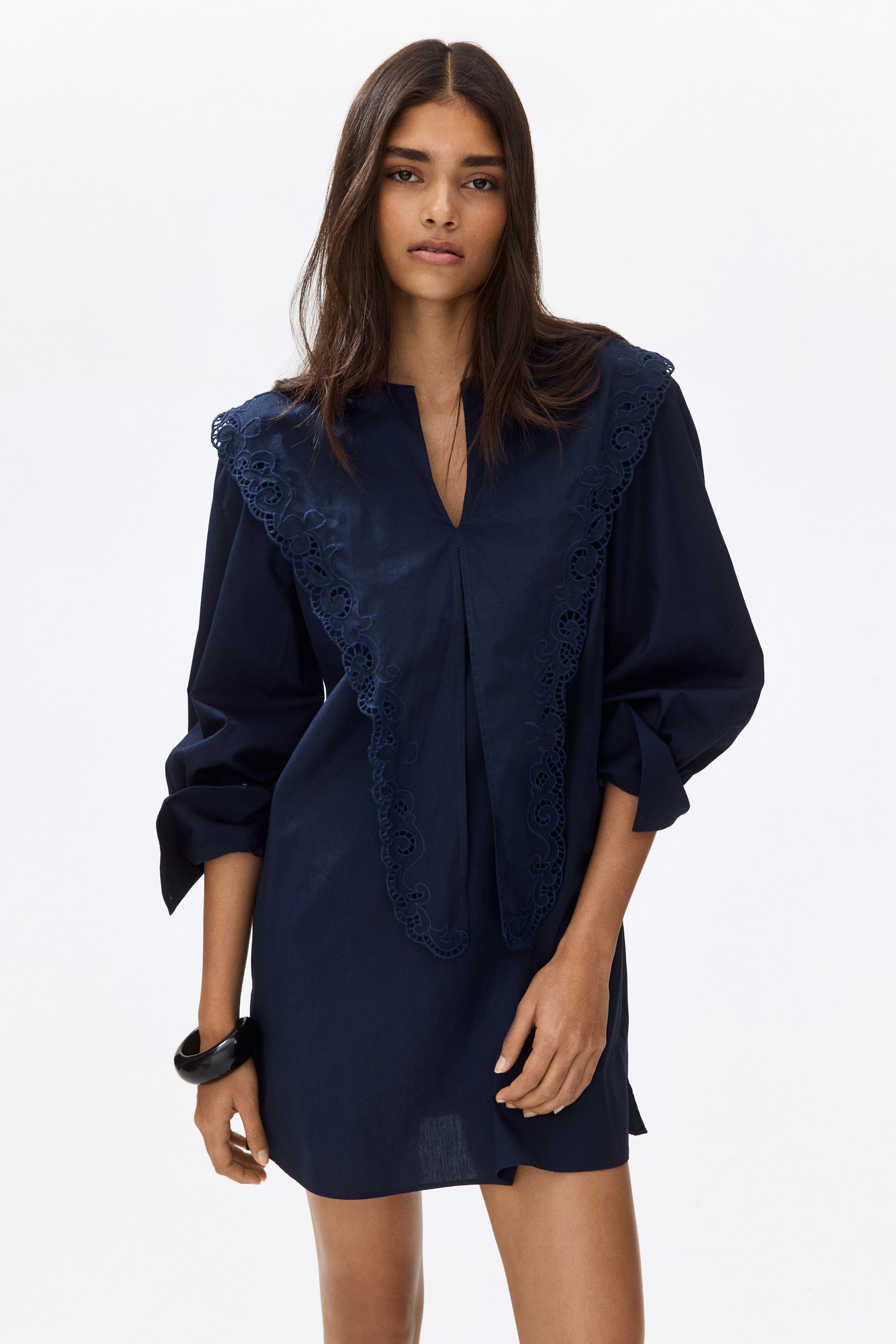 View larger image: A navy blue mini dress showcasing a deep V-neckline and a prominent, rounded collar adorned with intricate eyelet embroidery extending over the shoulders. The dress offers a relaxed fit with full-length sleeves rolled up to the elbows.