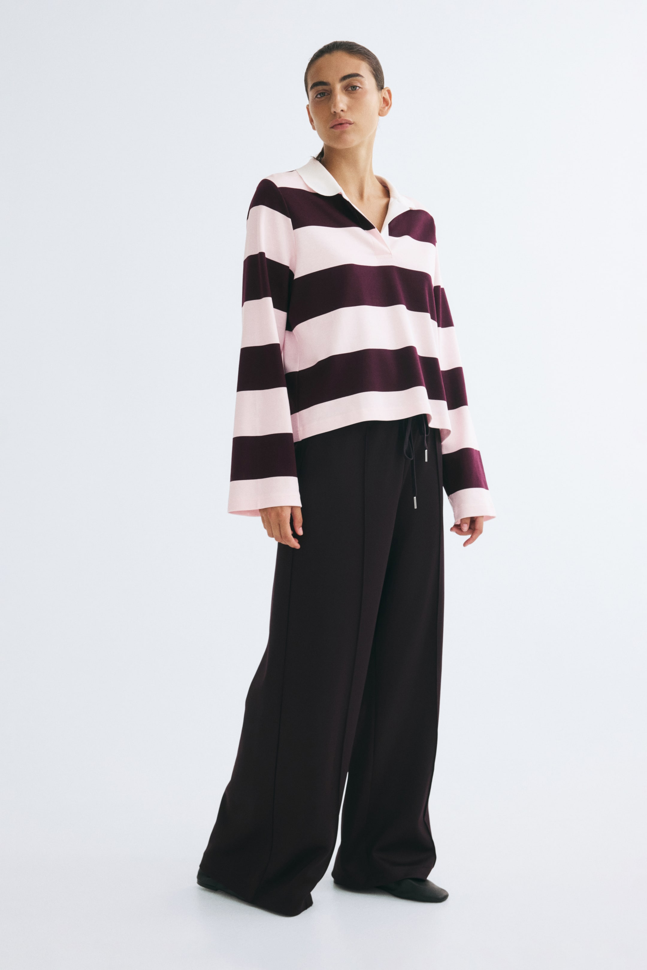 Ver imagen más grande: Esta polo a rayas en rosa claro y burdeos oscuro la lleva la modelo, y presenta un cuello abierto, mangas largas y anchas y un bajo recto. La camisa se combina con un pantalón oscuro de pernera ancha con cordón.