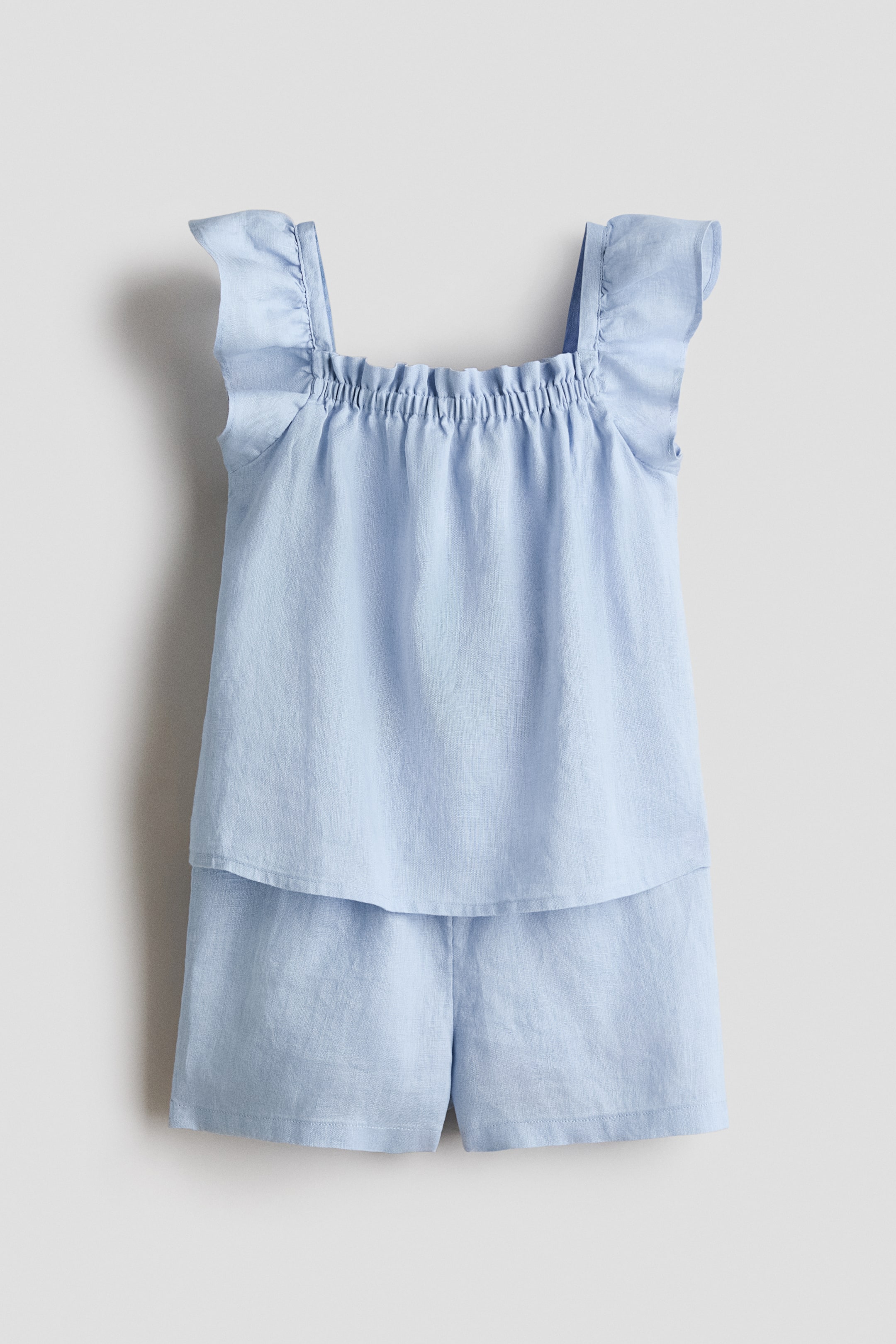 View larger image: A light blue two-piece set is displayed from the front, showcasing a top with a square, gathered elasticized neckline and ruffled shoulder straps. The top drapes loosely over the coordinating shorts, which are crafted with a relaxed fit.
