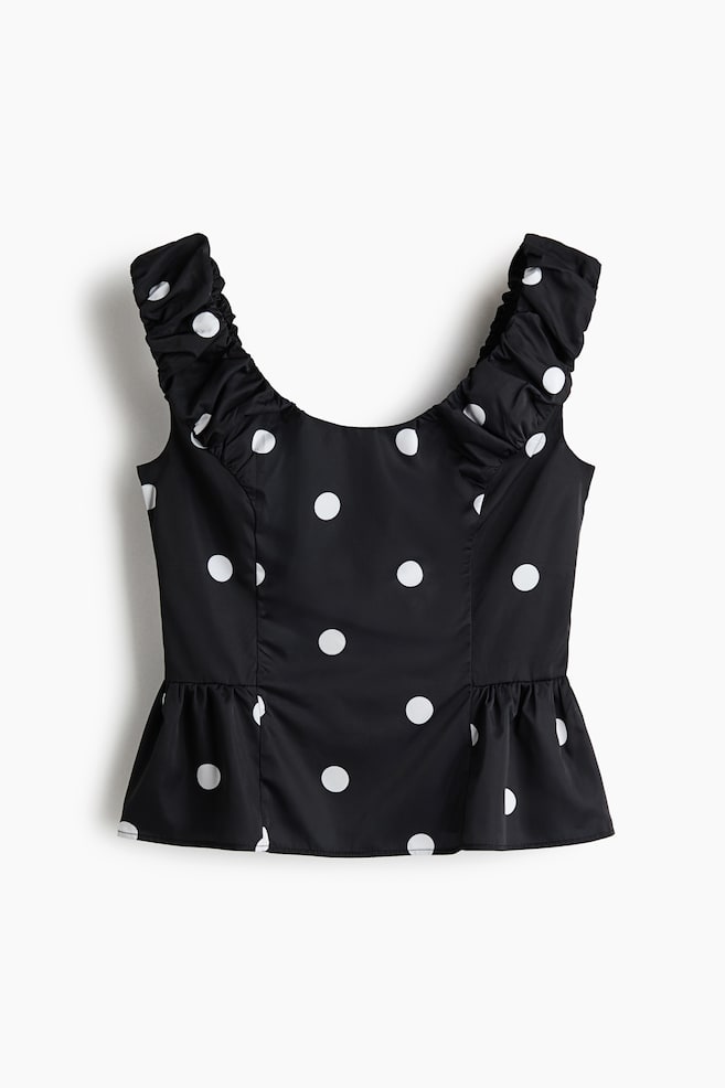 A black sleeveless peplum top featuring a white polka dot pattern is shown from the front. The top is designed with ruffled shoulder straps, a deep scoop neckline, and a gathered, flared hem. - 1