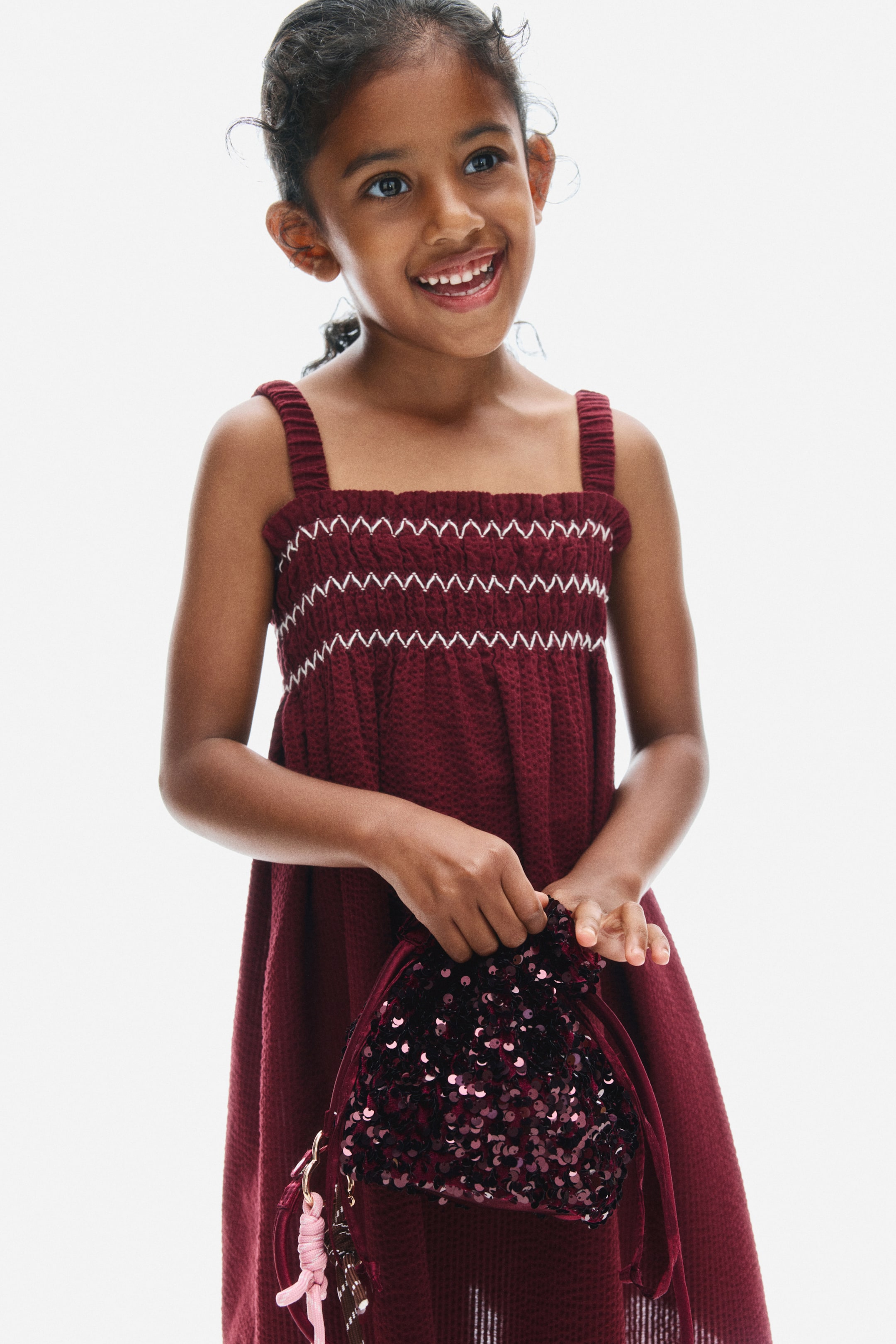View larger image: A dark red seersucker dress is shown from the front, featuring a smocked bodice with white zig-zag embroidery, a square neckline with a small frill trim, elasticated shoulder straps, and a flared skirt.
