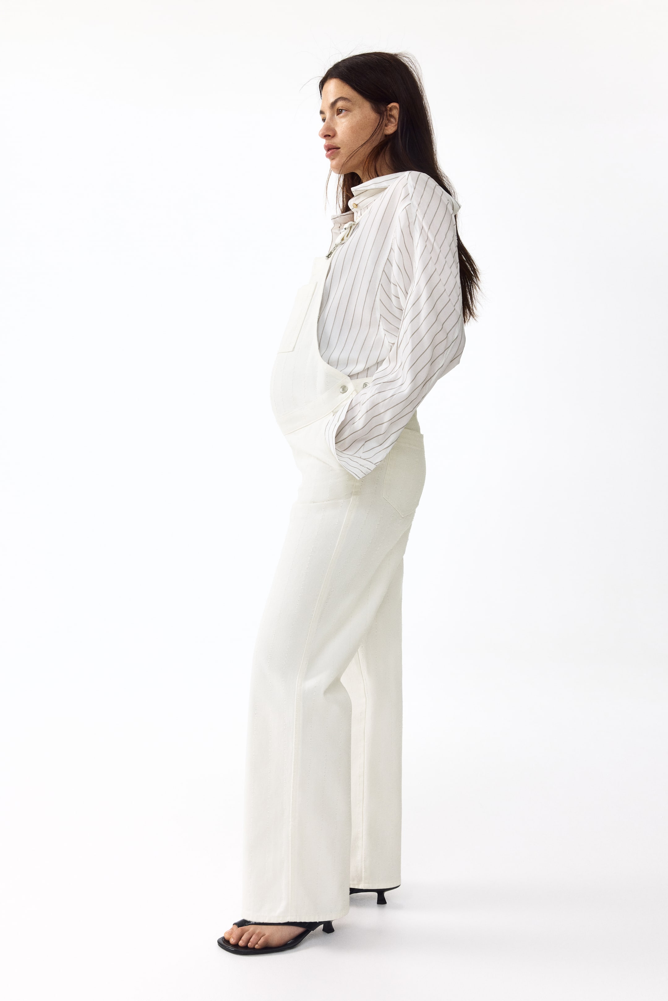 View larger image: Light off-white denim maternity overalls are displayed in a side profile, highlighting a front bib, side buttons at the waist, and wide-leg pants over a striped shirt.