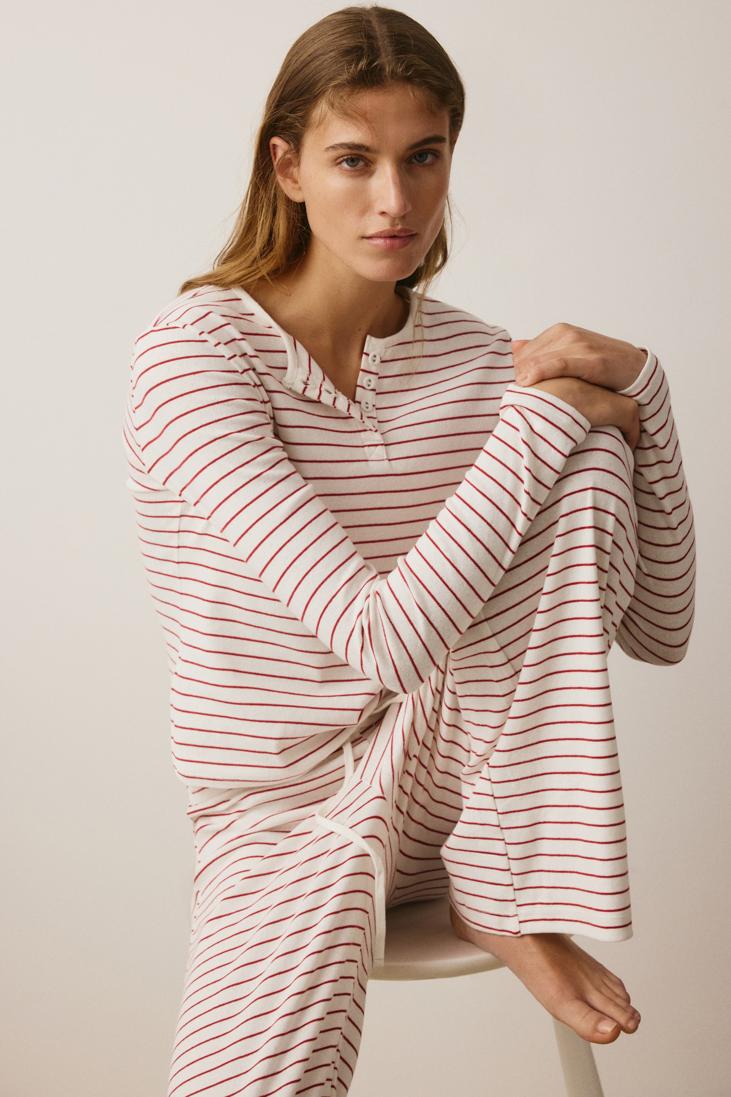 Ein zweiteiliges Lounge-Set ist in der Vorderansicht aufgehÃ¤ngt und besteht aus einem langÃ¤rmeligen Henley-Top und einer dazu passenden Wide-Leg-Hose. Beide Teile bestehen aus einem cremeweiÃen, gerippten Stoff mit dÃ¼nnen, horizontalen roten Streifen, wobei das Oberteil am Halsausschnitt eine kurze Knopfleiste hat.