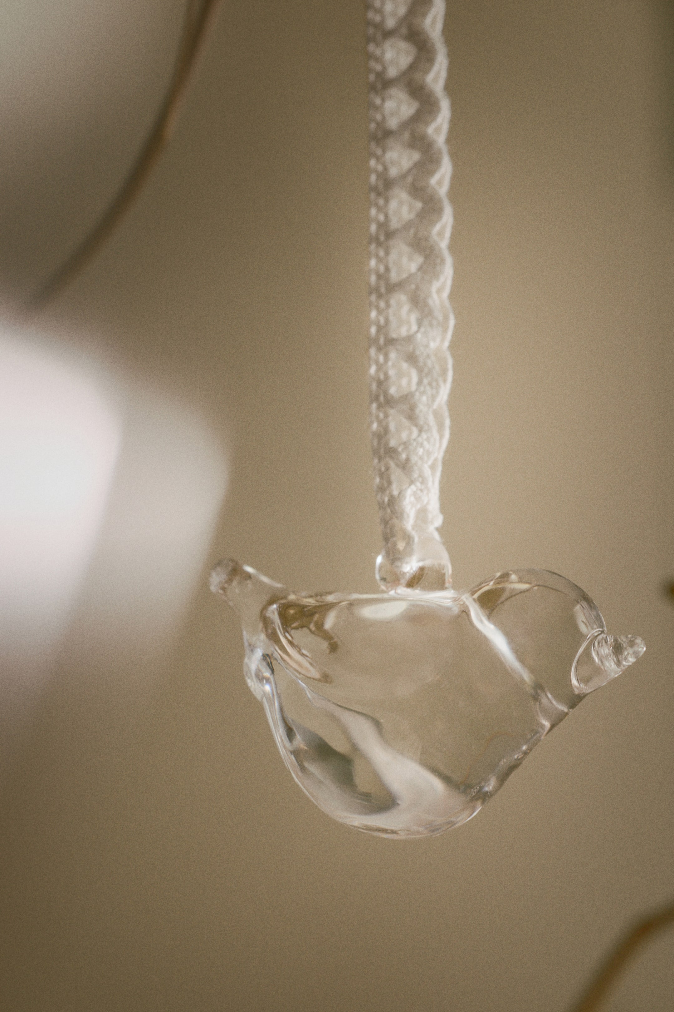 Grotere afbeelding bekijken: Een heldere glazen vogelornament, met een ronde lichaam, kleine snavel en staart, hangt iets schuin naar beneden en naar rechts aan een wit kantachtig lint met een opvallend geschulpte randpatroon.