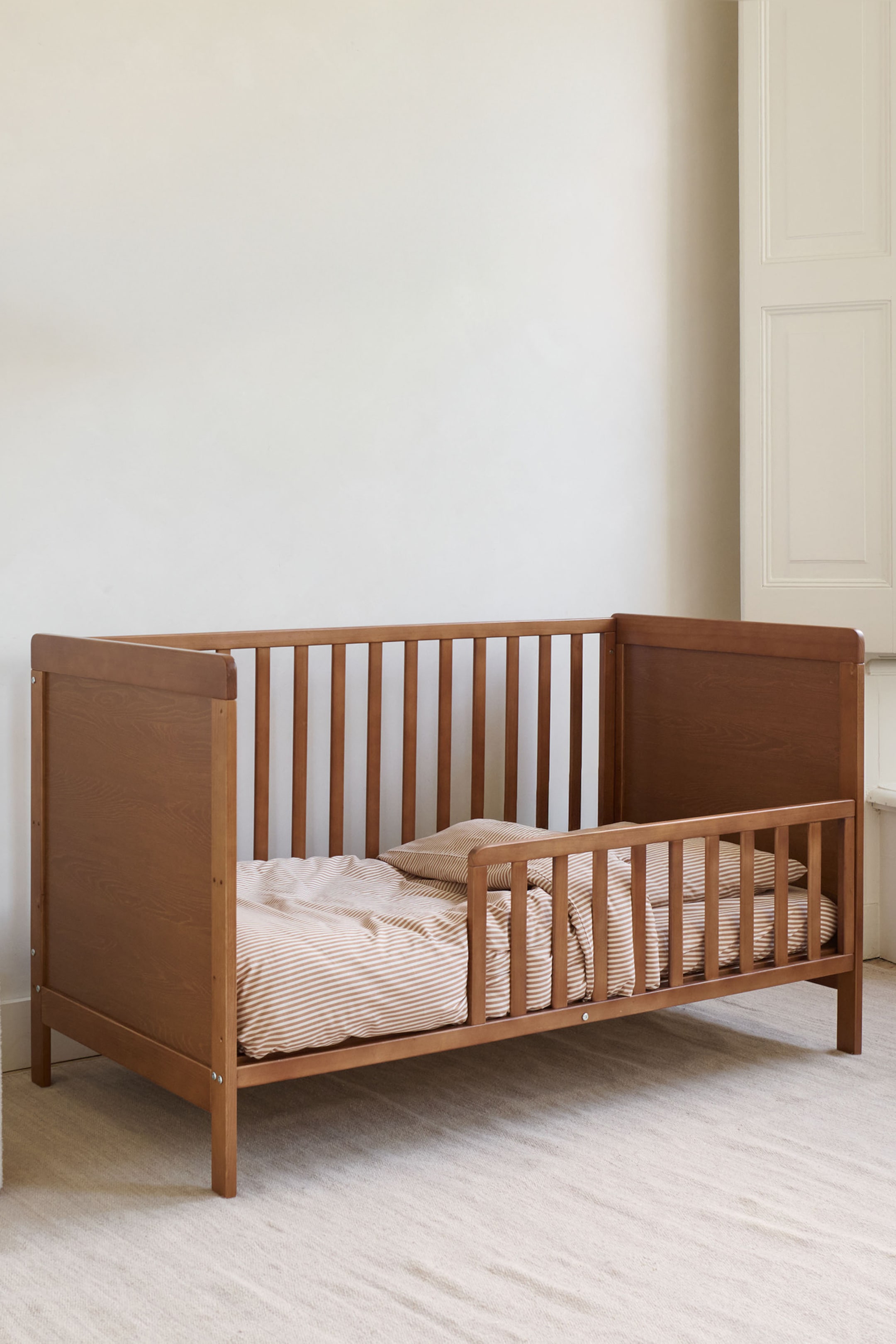 Agrandir l'image: Un lit pour tout-petit en bois présente une finition en bois brun clair avec des côtés à lattes verticales et des panneaux pleins pour la tête et le pied de lit, positionnés à un léger angle. Il est équipé d'un matelas, d'un oreiller et d'une couette, le tout recouvert de linge de lit rayé beige clair et marron.