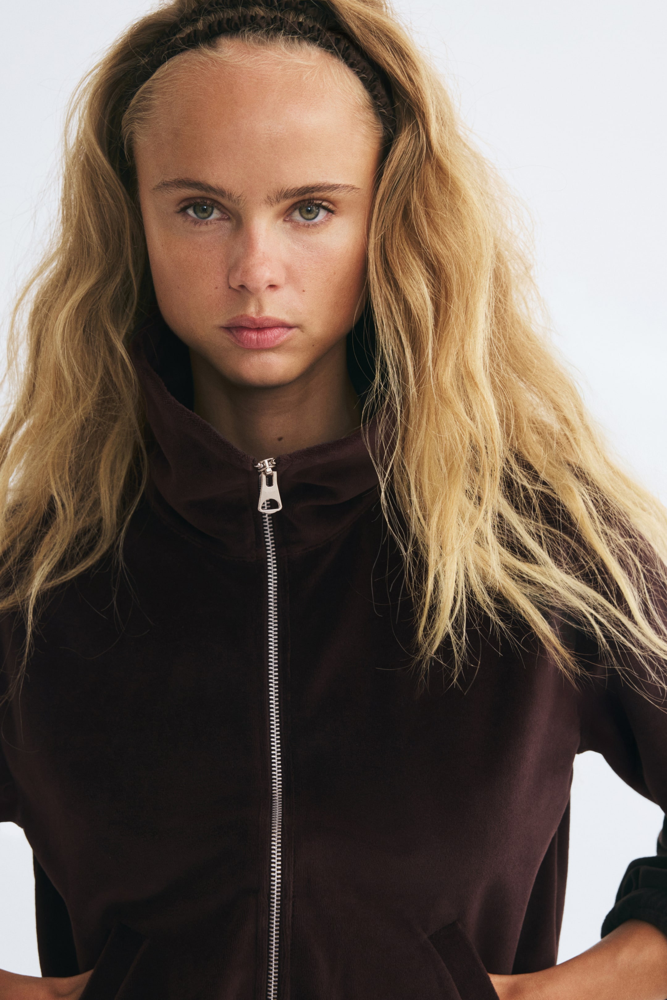 Visualizza l'immagine ingrandita: La modella indossa una giacca sportiva in velour marrone scuro, caratterizzata da un colletto alla coreana a doppio strato e da una zip frontale in metallo argentato. La modella posa con le mani infilate nelle tasche laterali a filetto della giacca.