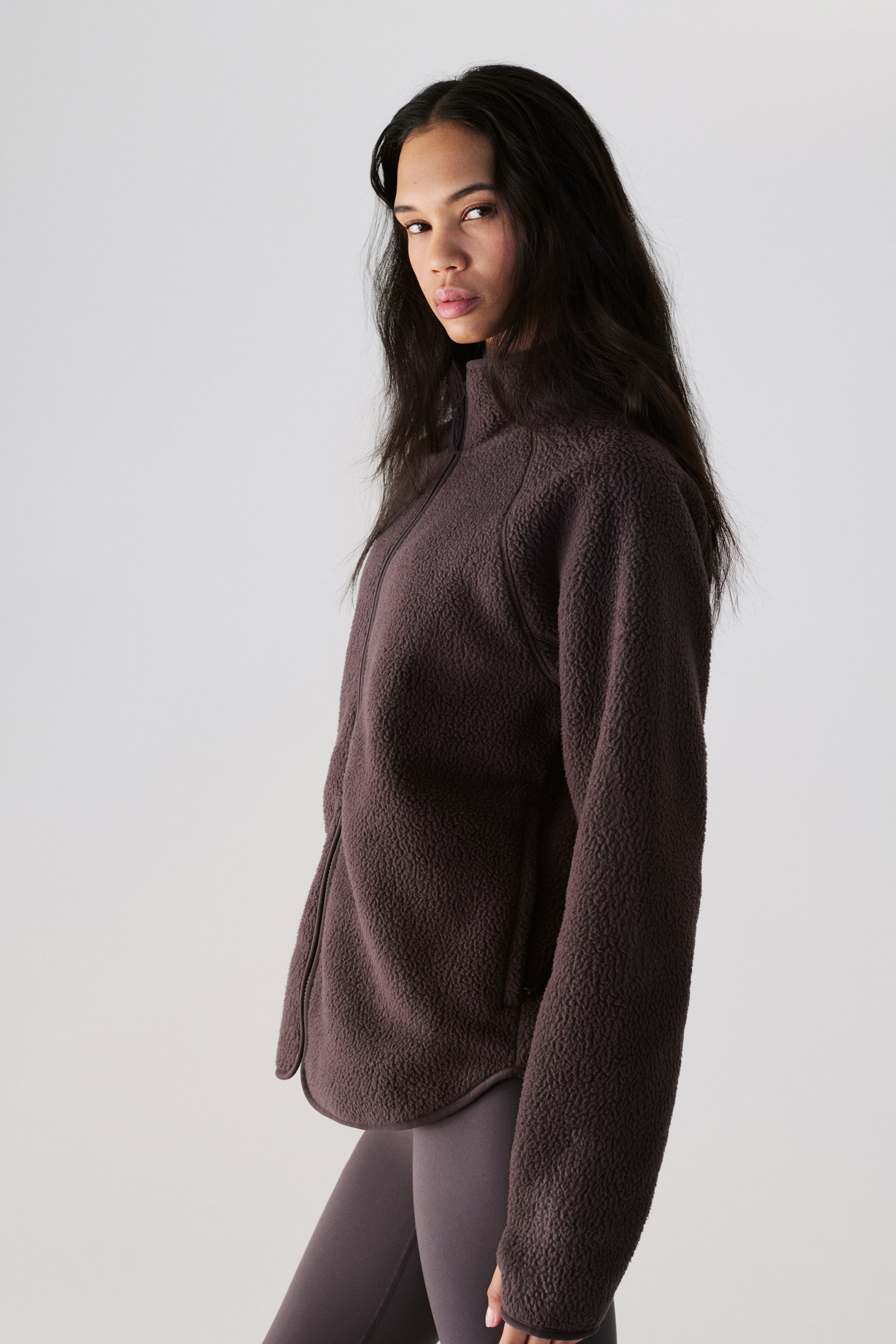 Nagyobb kép megtekintése: Egy sötétbarna texturált gyapjúdzsekit mutatunk be, amely magas gallérral, teljes hosszúságú elülső cipzárral, raglán ujjakkal és cipzáras oldalzsebekkel rendelkezik. Ívelt szegélyvonallal rendelkezik, és sötét leggings fölött viselik.