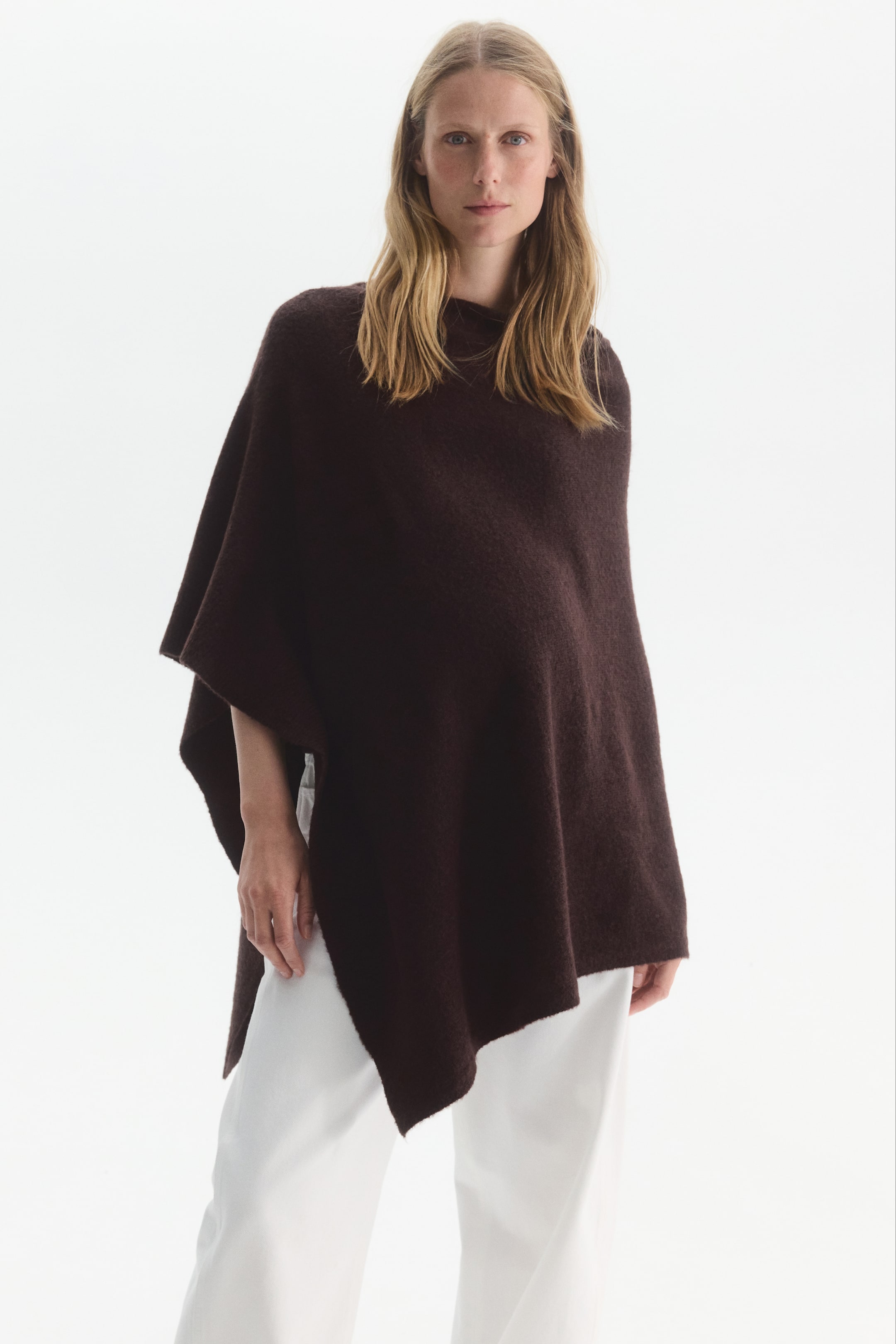 Grotere afbeelding bekijken: Een oversized, asymmetrische cape in een fijngebreide donkerbruine stof wordt door het model gedragen over een wijde witte broek. De cape heeft een boothals, een los vallend silhouet dat over een zwangere buik valt, en een ongelijke, puntige zoomlijn.