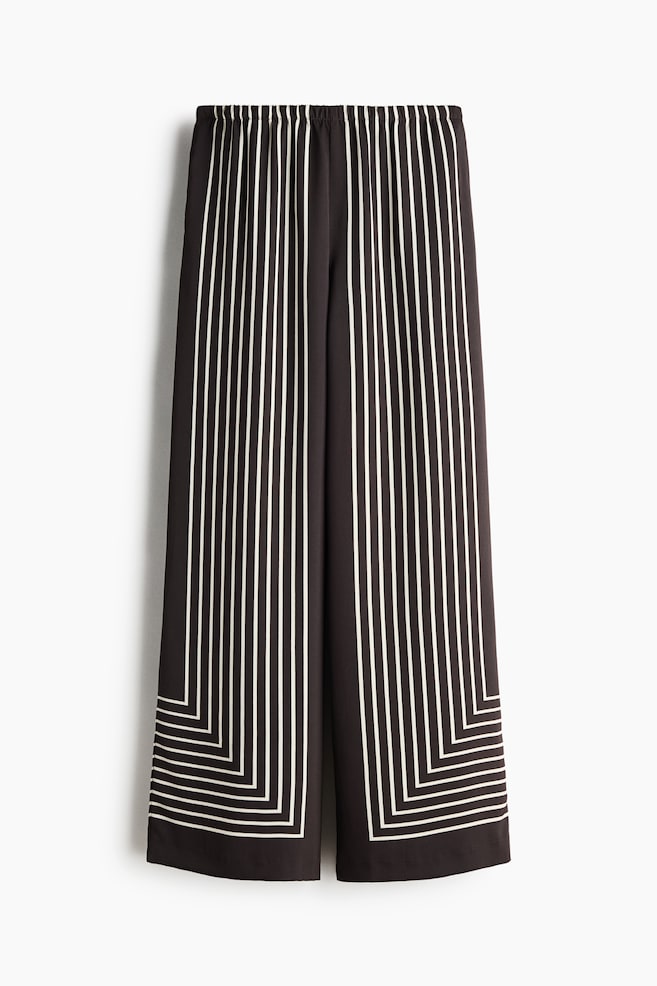 Von vorne zeigt die Hose eine hohe Taille mit verdecktem Gummizug und geraden Beinen. Sie ist aus dunklem Webstoff mit leichtem Glanz gefertigt. Weiße vertikale Streifen verlaufen die Beine hinunter und bilden im unteren Bereich jedes Beins ein geometrisches Quadratmuster. - 1