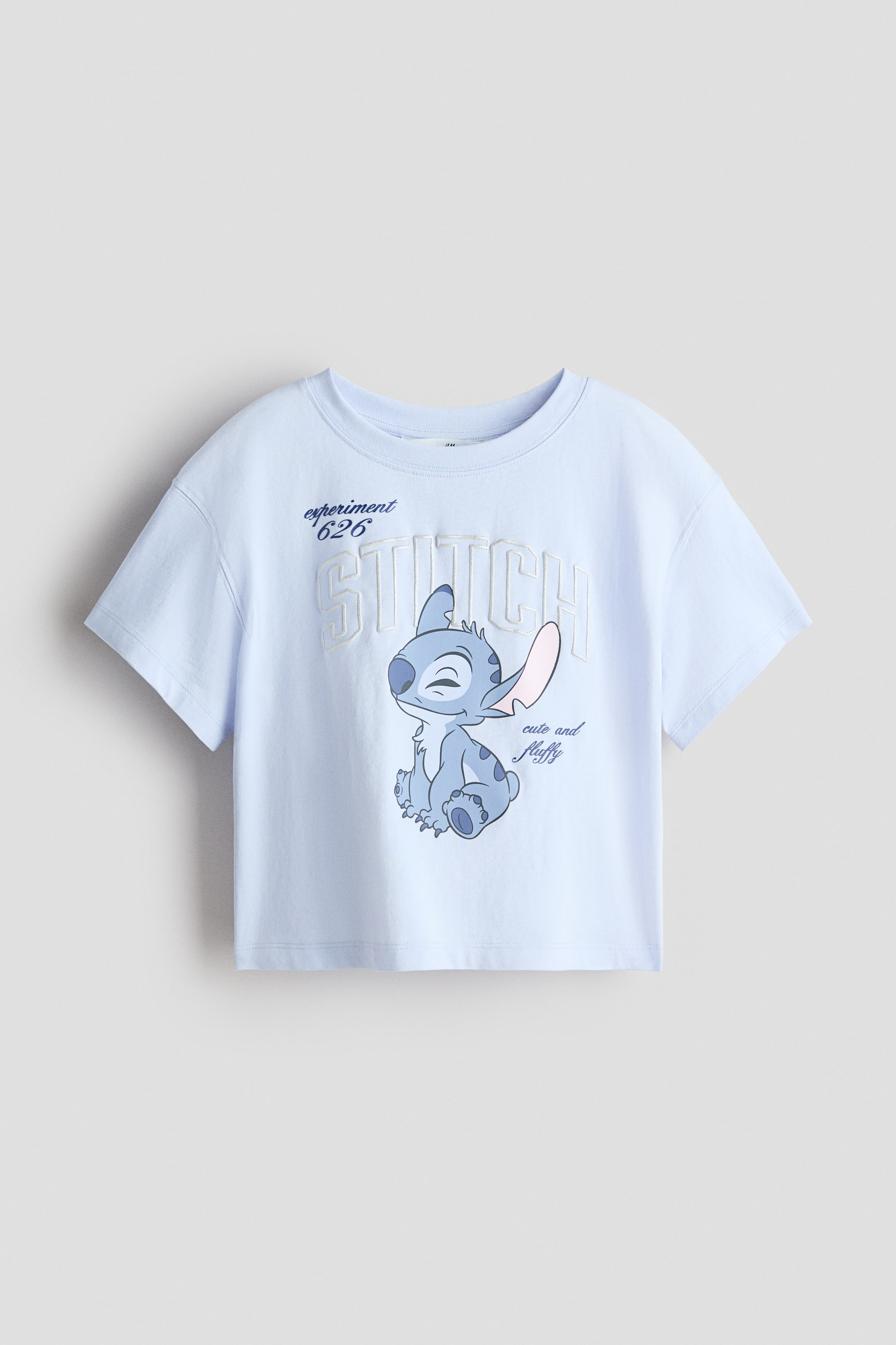 View larger image: A light-blue soft cotton-jersey top is shown from the FRONT, featuring a print motif of Stitch, "experiment 626," "STITCH," and "cute and fluffy" text. It is designed with a round, rib-trimmed neckline, dropped shoulders, and short sleeves.