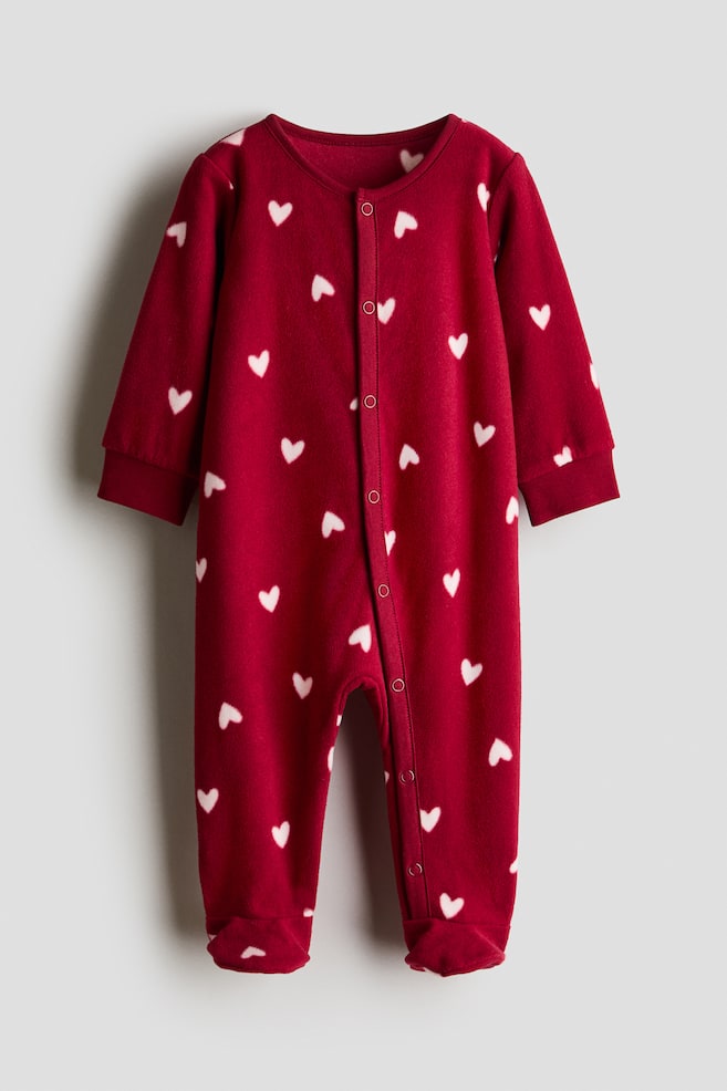 A red fleece fabric sleepsuit featuring an all-over white heart pattern is shown from the front. The sleepsuit features long sleeves with ribbed cuffs, footed legs, and a snap-button fastening running down the front. - 1